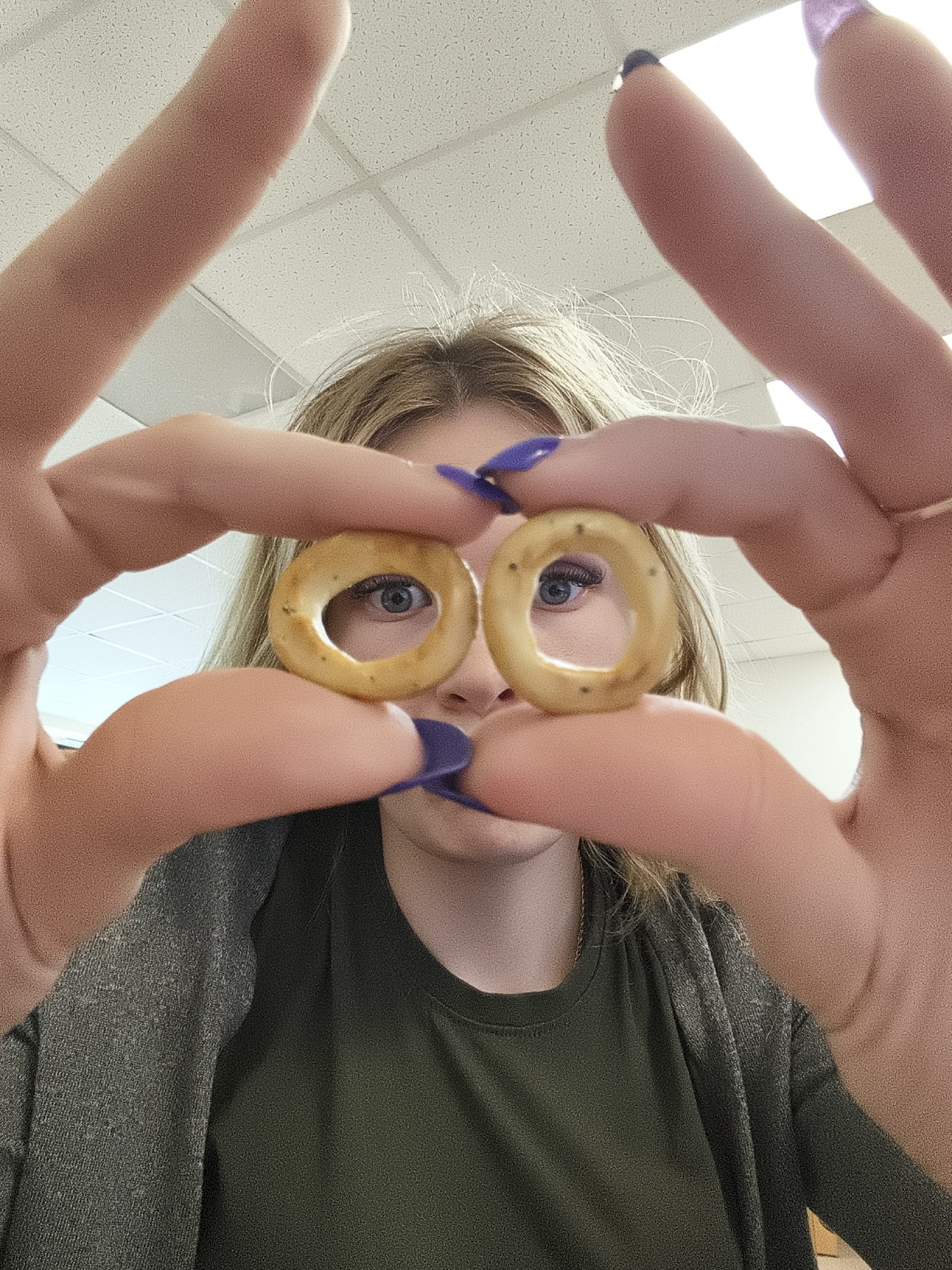 A young person is holding two small, circular, seeded bread rings, approximately the size of a quarter, up to their eyes, so that their eyes are visible through the holes in the rings. The person has light skin, light brown hair, and is wearing a dark green shirt and a grey cardigan. Their fingernails are painted purple. The background is blurry and indistinct, but appears to be a room with fluorescent lights. The location and city are unknown.