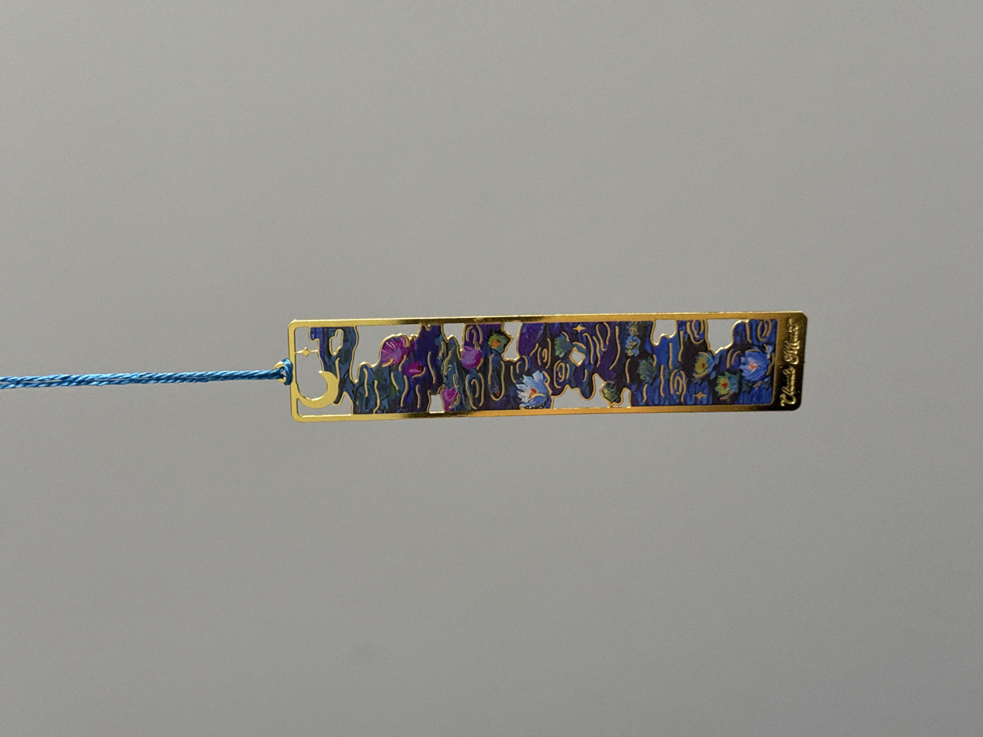 This is a close-up, eye-level shot of a gold-colored bookmark with an intricate, cut-out design featuring a floral pattern. The bookmark is horizontal and placed in the center of the frame. A thin, blue string is attached to the left side of the bookmark, extending horizontally to the left edge of the image. The bookmark itself has a golden border, and the interior is filled with a dark blue background adorned with colorful, abstract floral shapes in shades of purple, pink, and blue, with small accents of white. The artwork appears to be enameled or lacquered, giving it a glossy finish. On the right end of the bookmark, faint, etched text is visible, though it is not clearly legible. The background is a plain, neutral gray, providing no specific location cues beyond suggesting a studio or controlled environment for product photography. There are no people or animals present. The overall mood is neutral and focused on showcasing the detail of the bookmark.