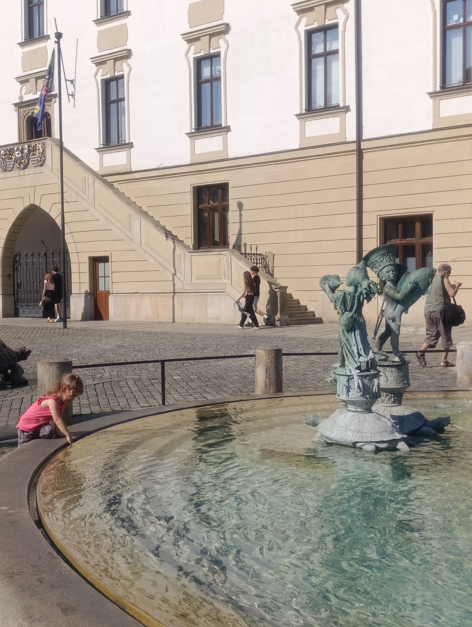 A young girl with blonde hair, wearing a pink tank top and patterned shorts, kneels at the edge of a fountain in Olomouc, Czechia. Her left hand is extended towards the water. The fountain features a bronze sculpture of two cherubic figures and several turtles. The fountain is surrounded by a stone border and cobblestone paving. In the background, a light-colored building with beige trim and several windows stands tall. A staircase leads up to a balcony on the building, where a flag is displayed. Several people are walking in the background, and shadows are cast on the ground, indicating it is daytime and sunny.