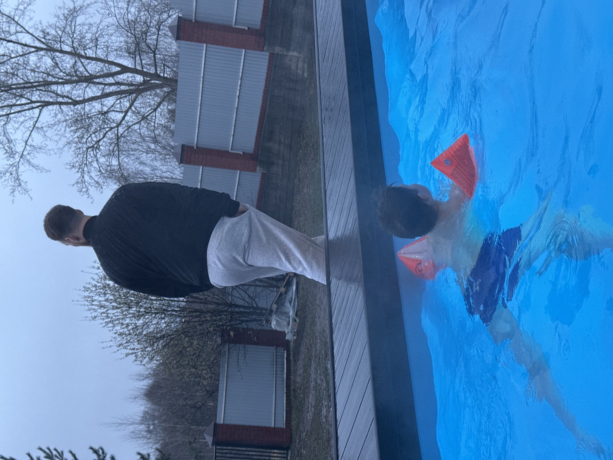 A man wearing a black jacket and light-colored pants stands beside a pool. A child is swimming in the pool with orange inflatable armbands. The pool has a dark blue lining. A fence is visible in the background, along with trees. The photo was taken in the city of Perm, Russia.