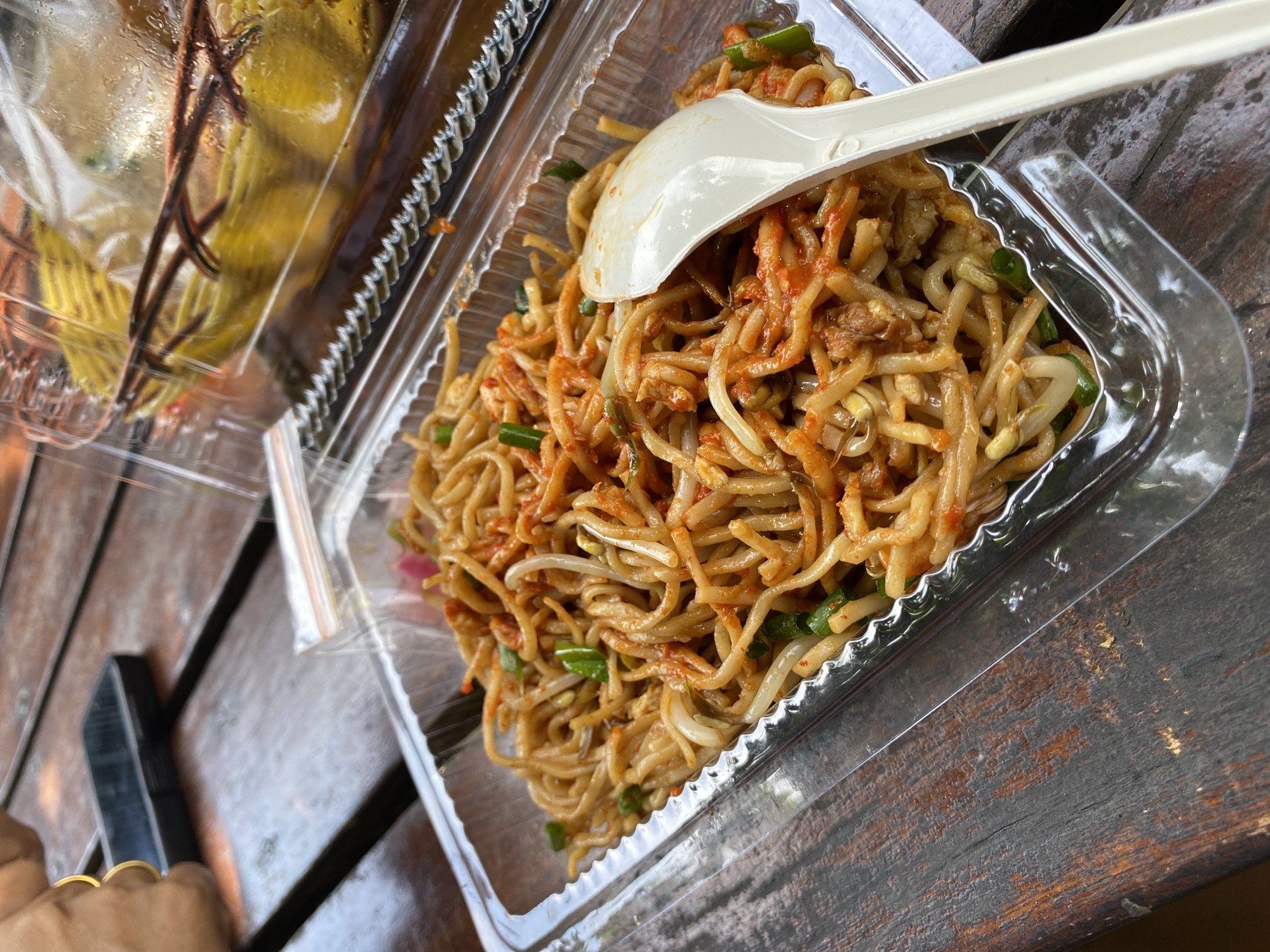 In Bireuenmeunasahdayah, Indonesia, a container of stir-fried noodles sits on a weathered wooden table. The noodles are tangled and coated in a reddish sauce, interspersed with green onions and bean sprouts. A white plastic spoon rests partially in the noodles, positioned to the right of the frame. In the background, a clear plastic bag contains what appears to be yellow food items, possibly fruits or pastries, contained within a woven basket. A portion of a person's hand, adorned with a gold ring, is visible in the upper left corner, along with a black mobile phone lying on the table. The lighting suggests an outdoor setting, possibly a cafe or food stall.