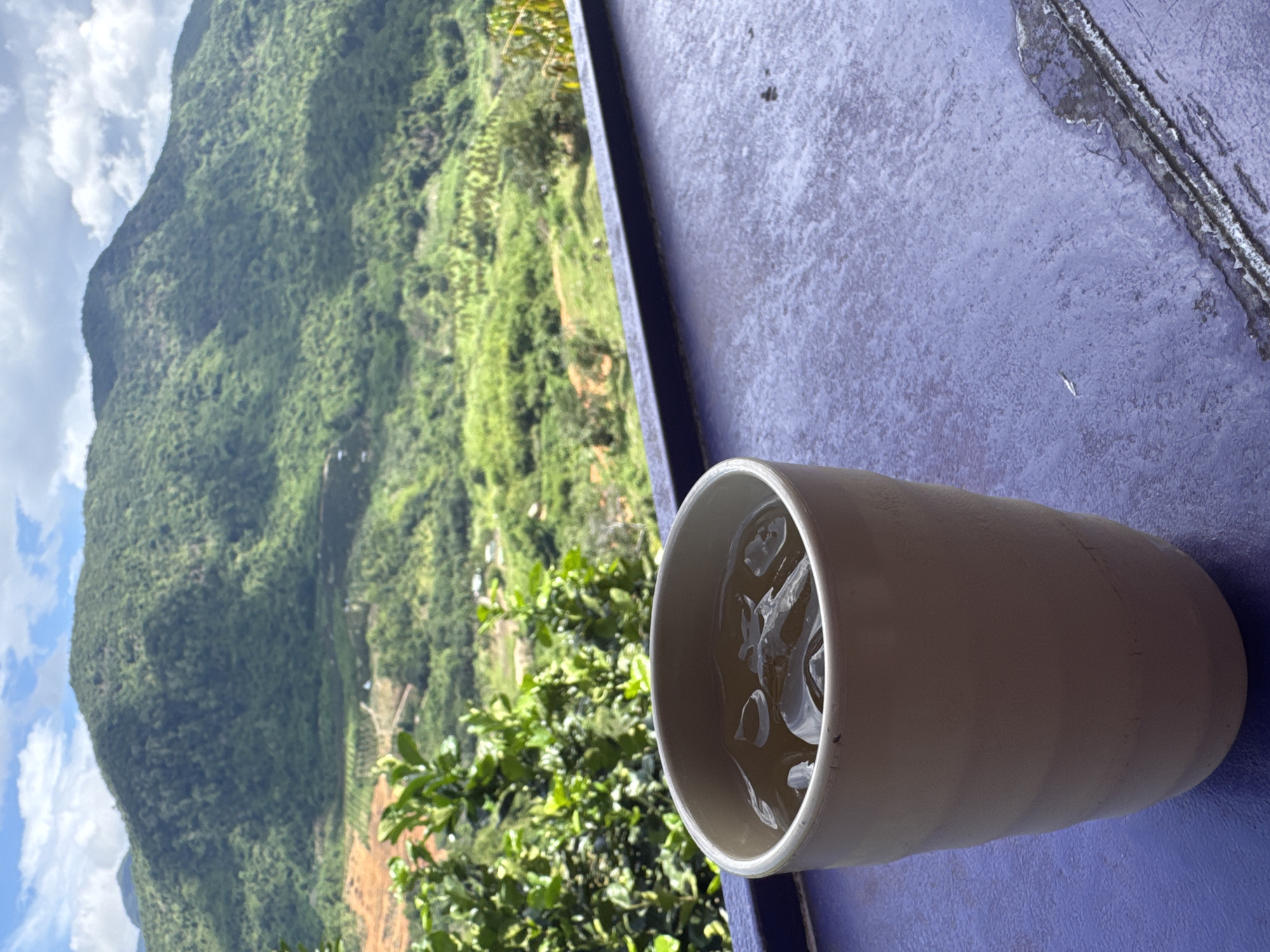 A close-up shot features a light-colored ceramic cup filled with ice and a light-colored beverage. The cup sits on a textured purple surface. In the background, lush green mountains with some terraced fields and scattered structures dominate the view under a cloudy sky. The overall scene suggests a tranquil, possibly rural, setting.