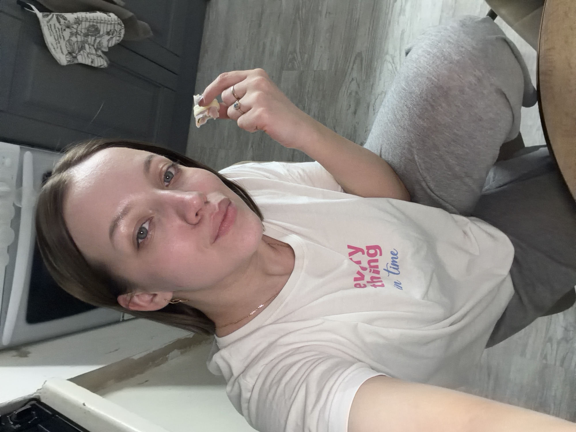 A woman is seated indoors, holding a piece of food between her fingers, possibly about to eat it. She is wearing a white t-shirt with writing on it and grey sweatpants. The background features kitchen cabinets and flooring. The specific location is Reshetnikovo, Russia.
