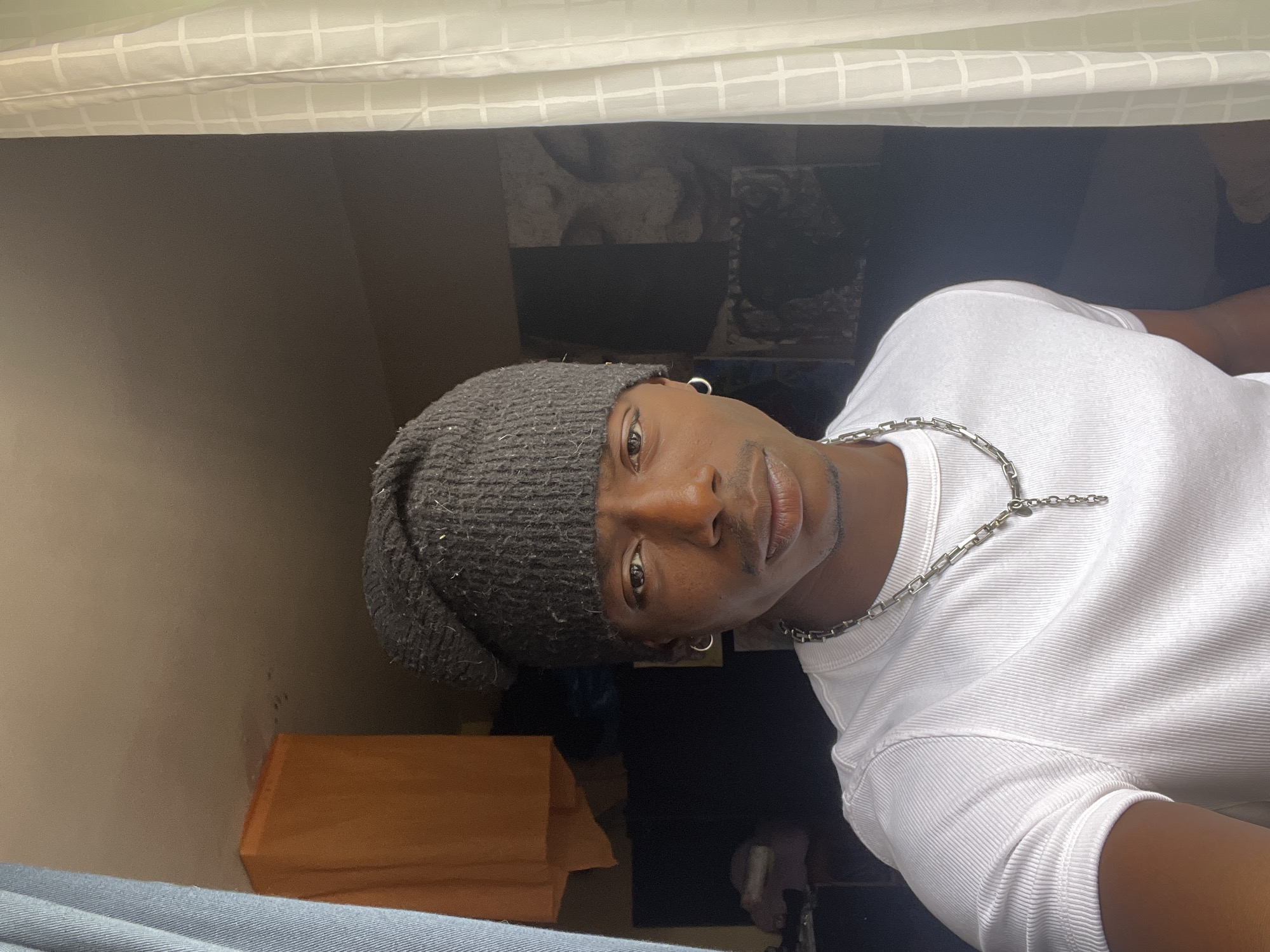 A medium indoor shot of a person with brown skin wearing a grey beanie, silver hoop earrings, and a silver chain necklace, and a white ribbed shirt. They are positioned at an angle, looking directly at the camera. The background includes a wooden shelf, a dark window, and a white curtain or sheet. The person appears to be indoors, possibly in a bedroom or living space.
There is no visual information to suggest the location is Barcelona, Spain.