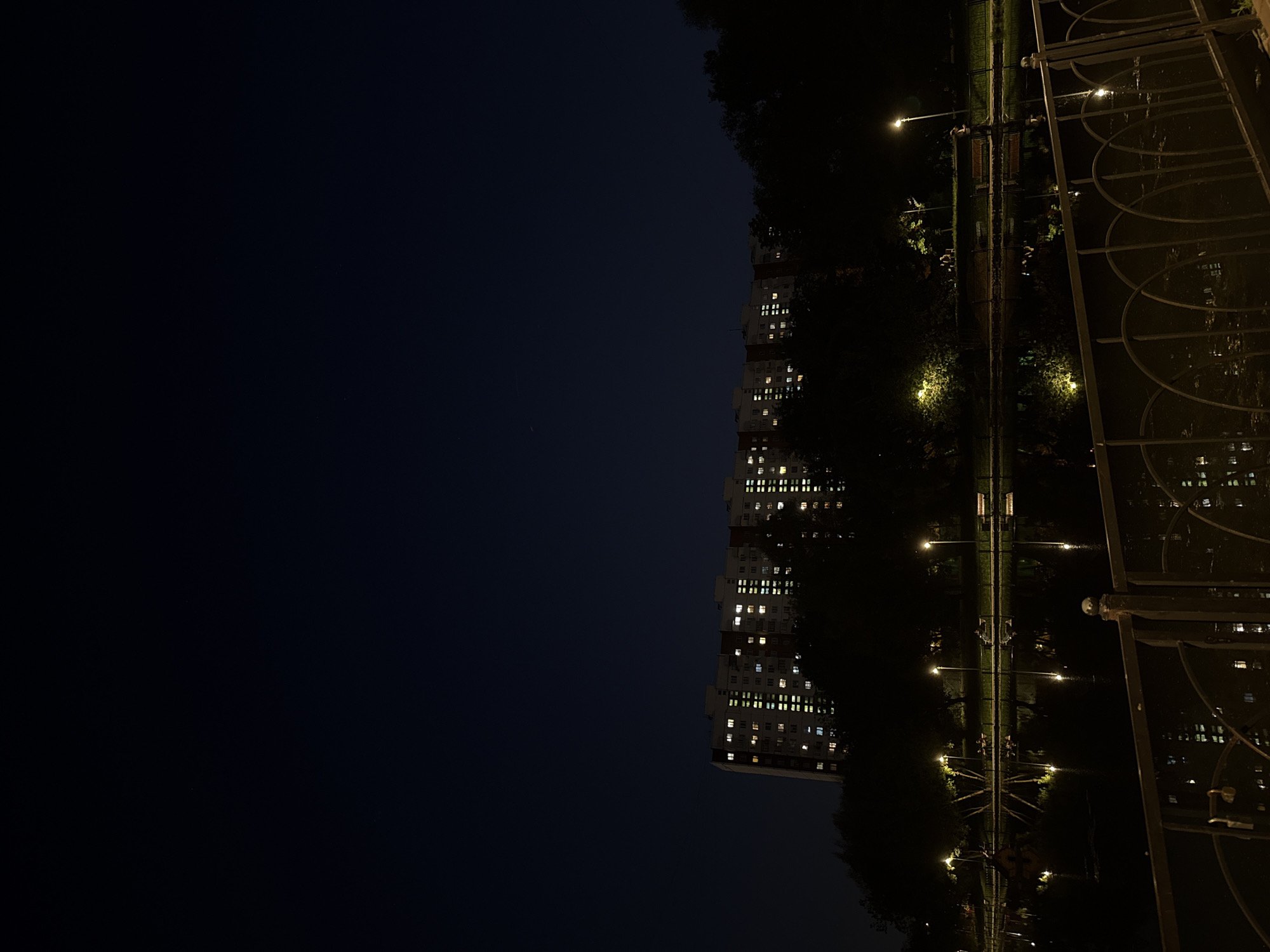 The image shows a nighttime view of a city, likely Moscow, Russia. A tall apartment building with illuminated windows is on the left. Streetlights illuminate a park pathway with benches and trees. A decorative metal fence is in the foreground on the right. The dark sky is visible on the left, and the overall scene suggests a quiet urban night.