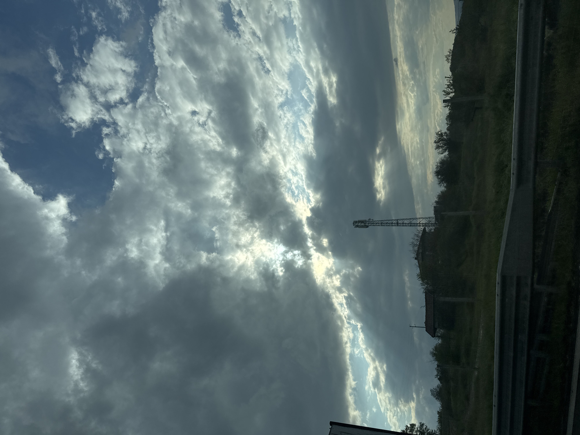 The image, currently oriented sideways, captures a striking daytime landscape under a dramatic sky, likely viewed from a moving vehicle or roadside in Zvezditsa, Bulgaria. The upper two-thirds of the frame are dominated by a dynamic sky featuring a mix of dark, heavy grey clouds and brilliant patches of sunlight. Sunbeams penetrate the cloud cover, creating visible crepuscular rays that illuminate parts of the atmosphere, while sections of clear blue sky are also visible in the upper left.
Below the tumultuous sky, a verdant, hilly landscape extends to the horizon, covered with lush green vegetation, including numerous trees and bushes. A tall, slender metal lattice tower, characteristic of a telecommunications or radio mast, stands prominently on the right side, silhouetted against the bright sky. Further below the tower, partially hidden by foliage, is the dark outline of a building with a distinct roofline.
A sturdy metallic guardrail runs vertically along the right edge of the image (horizontally if viewed correctly), indicating proximity to a road or highway. A sliver of a white structure is visible in the bottom right corner. The interplay of light and shadow suggests a day with shifting weather patterns, possibly late afternoon or early morning. There are no people, animals, or specific events taking place. No visible text is present.
