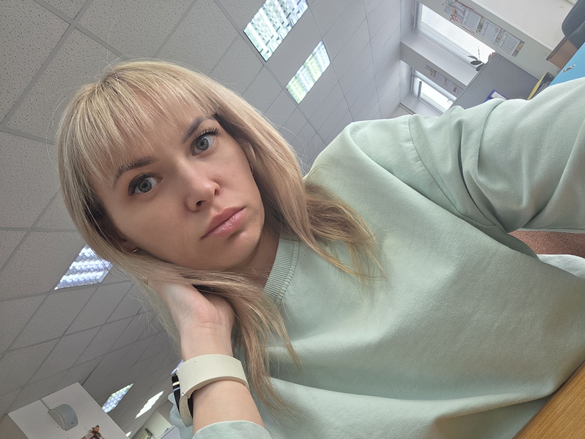 This media file presents a low-angle, close-up photograph of a woman in an indoor setting, likely an office in Chaikovsky, Russia. The subject is a woman with blonde hair, styled with bangs, and light-colored eyes. She is looking directly at the camera with a somewhat pensive or thoughtful expression, leaning her head on her left hand. She is wearing a light mint green crewneck sweatshirt and a white or light-colored smartwatch on her left wrist.
The background reveals typical office architecture: a drop ceiling with a grid pattern and numerous recessed fluorescent light panels. Natural light appears to enter through windows with blinds visible in the upper right background. A wooden desk or table surface is partially visible on the right side of the image. Further in the background, there are hints of office furniture, including what might be part of a dark blue office chair and an indistinct item on a wall, possibly a calendar or poster, though no text is legible. The overall scene suggests a moment of quiet or reflection during the daytime in a professional work environment.