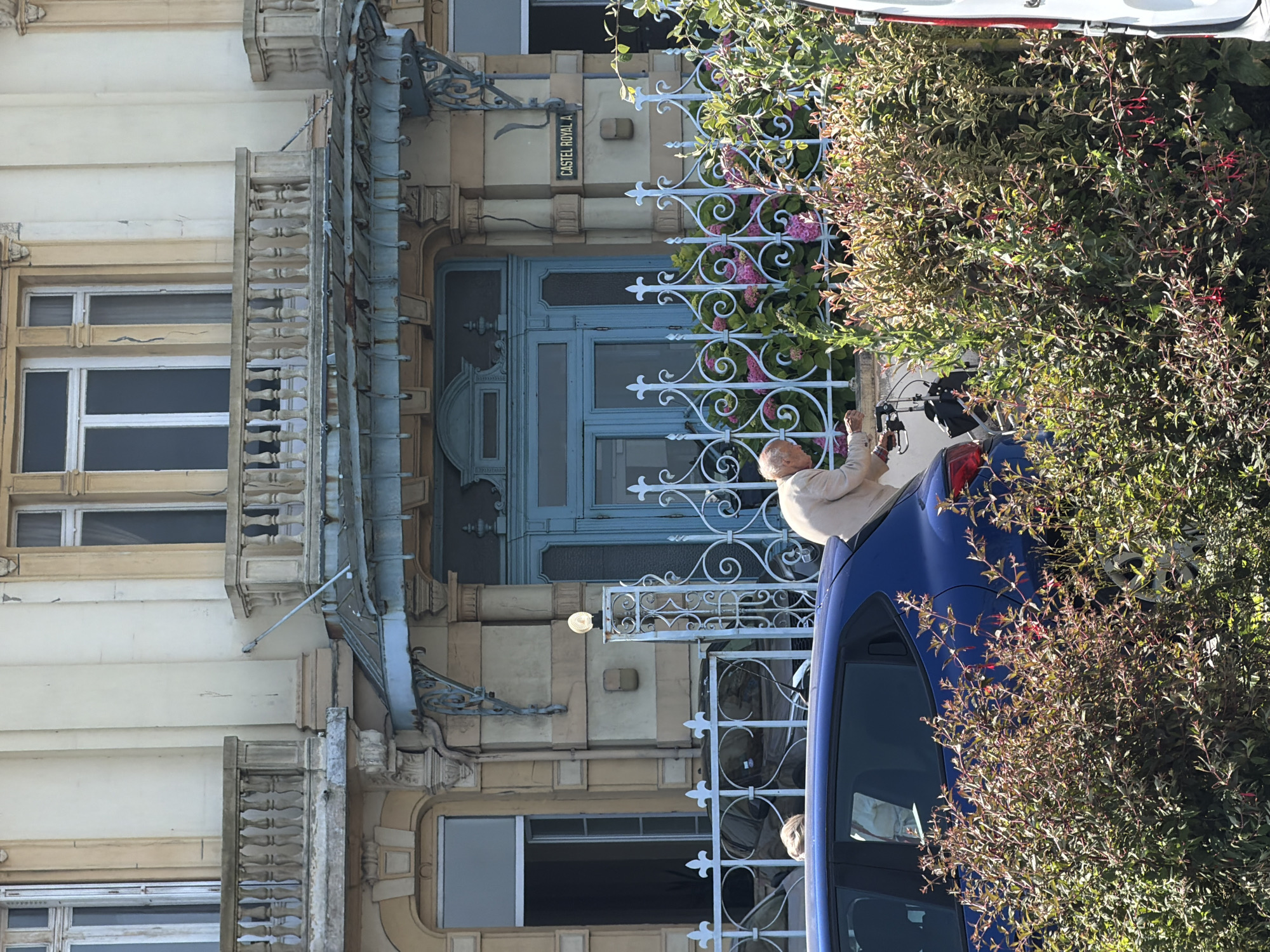 An elderly Caucasian man, wearing a light-colored jacket, is seen from the side, operating a walker in front of a blue vehicle. He is positioned near a white ornate fence with a flowering bush in front of it. Behind the fence, a light blue, ornate door is visible, with a sign that reads "CASTEL ROYAL A." The scene takes place in front of a building with balconies and large windows, characteristic of the architecture found in Dieppe, France.