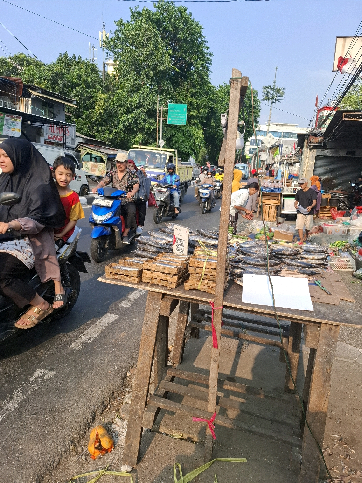 The image captures a street scene in South Jakarta, Indonesia, likely in a market area. The foreground features a wooden stall displaying numerous fish, some neatly arranged in wooden crates and others laid out on the ground. A blank white paper rests on the table, and to the right, small dried fish are scattered. A wooden post with red ribbons tied to it stands prominently.
In the mid-ground, a busy street with motorbikes and a truck is visible. A woman wearing a black hijab and a child in a red and yellow shirt are seated on a motorbike. Another motorbike with two adults is also present. Pedestrians are seen near the market stalls.
The background shows a street lined with buildings and trees, with a cell tower visible above the greenery. The sky is clear and blue, suggesting it is daytime. The overall impression is of a vibrant, bustling street market with commercial activity.