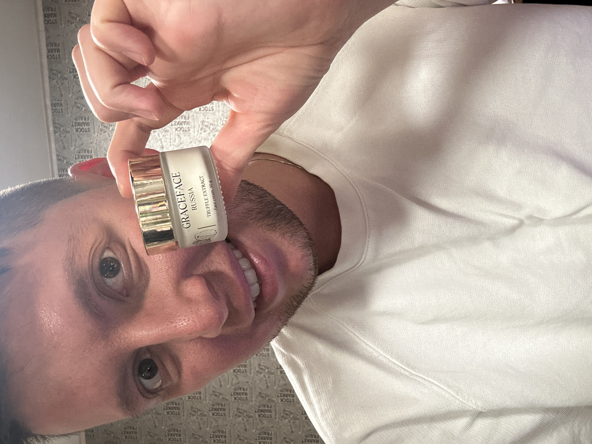 A man holds a small jar of "Graceface Russia Truffle Extract Face cream, 30g" close to his face. The background consists of a wall with a repeating pattern of the words "STOCK", "MARKET", and "FRAU." The man is wearing a white sweater. The location, based on the cream's label, is likely Vorotynsk, Russia.