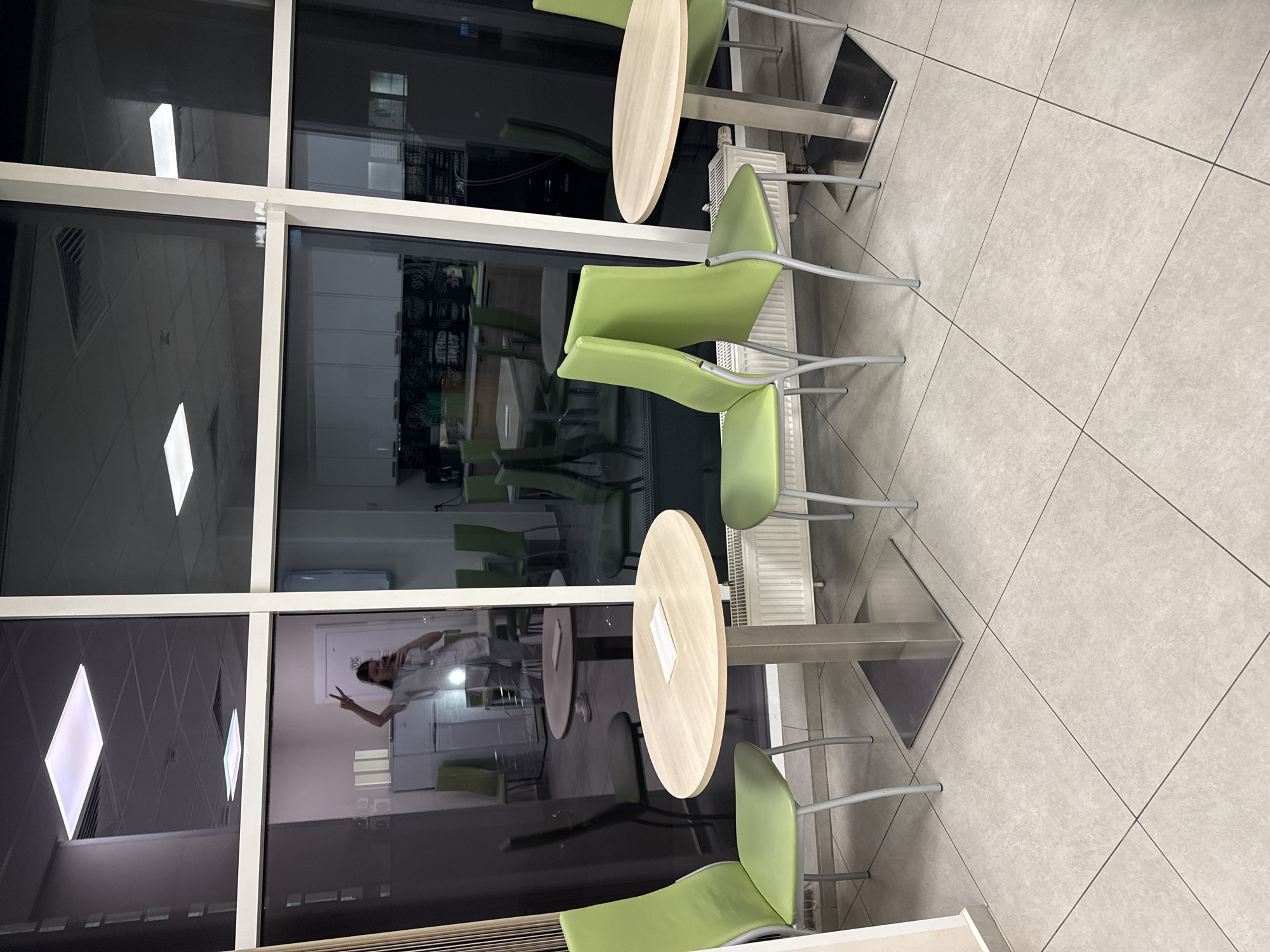 The image displays an interior space, likely a cafeteria or break room, featuring modern light-colored round tables with metallic bases and several lime-green chairs with metal frames. The floor is tiled with light gray squares separated by darker grout lines.
A significant portion of the scene is dominated by a wall of large glass windows or partitions, divided by white frames. These glass panels prominently reflect the indoor environment, including bright, square-shaped ceiling lights.
A person is clearly visible in reflection on the lower-left window pane. This individual, wearing a light-colored top, is holding a device, likely a smartphone, with their left hand as if taking a picture or selfie. With their right hand, they are making a "peace sign" (V-sign), conveying a cheerful or playful mood. Behind the reflected person, a white cabinet or refrigerator is also visible in the glass.
Other elements reflected in the windows include parts of a white radiator and what appears to be shelving or storage units in the background. On one of the tables, a white tissue or napkin is present. The artificial lighting throughout the space suggests it could be evening or a windowless area reliant on internal illumination. The room appears otherwise unoccupied, capturing a quiet moment. No explicit text is clearly legible within the image.