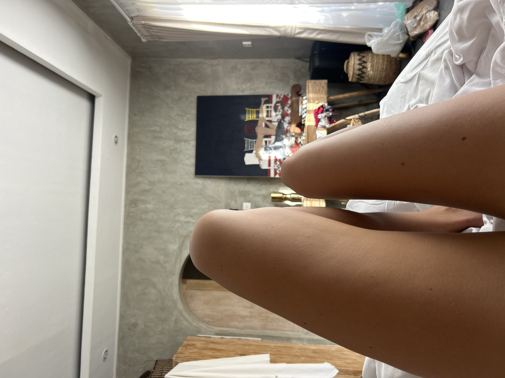 The image, originally rotated ninety degrees counter-clockwise, depicts an indoor scene with a person's bare, tanned legs occupying the foreground, partially covered by white bedding or fabric. The setting appears to be a modern room, possibly a bedroom or living area.
In the midground to the left, a white sliding door or wardrobe is visible. The room's walls are finished with a raw, textured gray concrete. A prominent feature on the main wall is a large rectangular painting with a dark background, showcasing stylized buildings and figures in red, white, and yellow. Below the painting, a wooden furniture piece and a distinctive circular cut-out are integrated into the concrete wall.
Towards the right side of the room, a woven basket and a black bag are visible, along with a golden fixture, possibly part of a lamp or plumbing. A wooden bench or low table with white paper or fabric rests in the lower right. The upper part of the image reveals window blinds or curtains, suggesting natural light is entering the room.
The scene portrays a moment of relaxation, with the individual likely resting or seated. The minimalist decor, concrete walls, and natural wood elements suggest a contemporary aesthetic common in resort or modern living spaces in North Kuta, Indonesia. The lighting indicates daytime. No visible text is present.