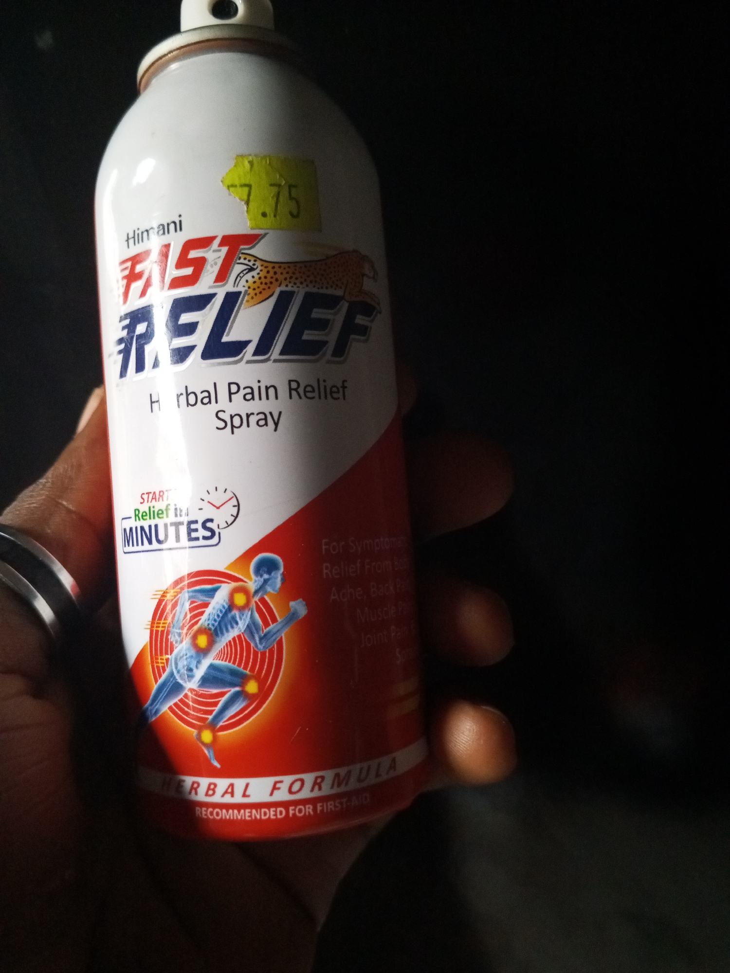 An overhead shot shows a hand holding a can of Himani Fast Relief Herbal Pain Relief Spray in Ashata, Nigeria. The can is white with red and blue lettering. A price tag is visible on the top left of the can, which reads "7.75". The can also features a graphic of a leopard and a diagram of the human skeletal system with highlighted joints. The background is dark and out of focus, with some light glinting off the can.