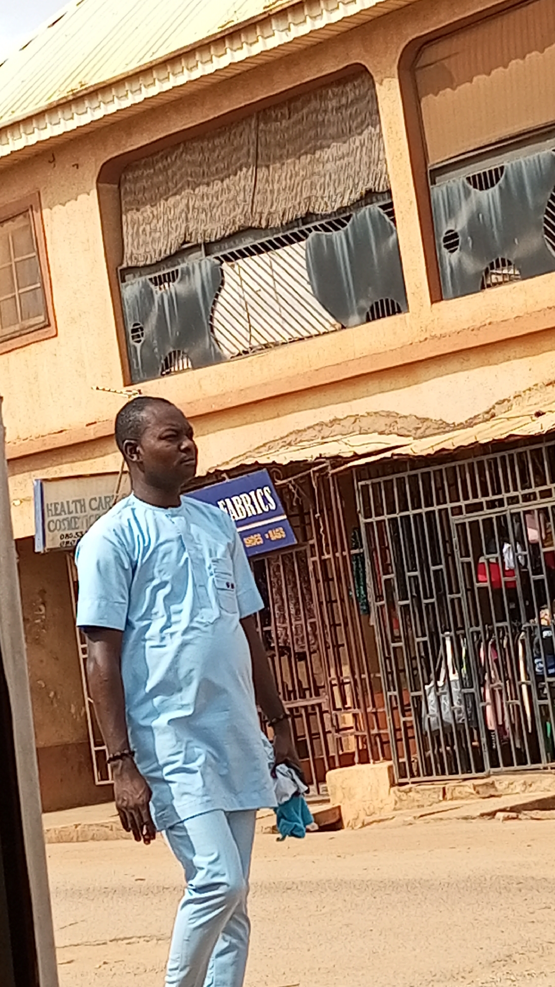 A man in a light blue traditional tunic and pants walks on a street. He has dark skin, a shaved head, and is wearing a bracelet on one wrist. He carries a crumpled blue and white cloth in one hand. Behind him are commercial buildings with metal bars and signs. A sign for "Fabrics" is visible, along with one for "Health Care Cosmetics." The buildings are painted in earthy tones. The location is in the city of Unknown, Unknown.