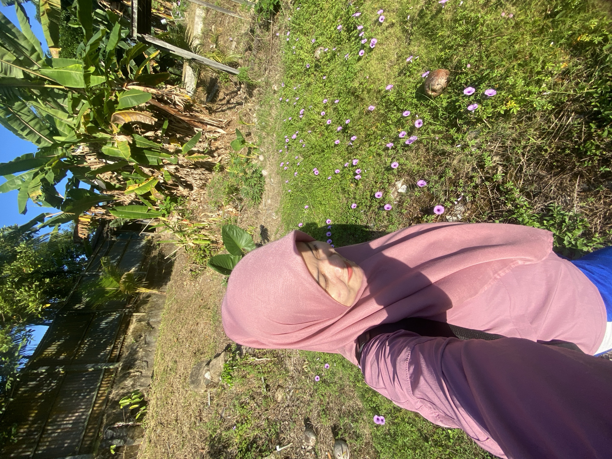 A selfie is taken by a woman wearing a pink hijab and a pink long-sleeved shirt. She is standing in a grassy area with small purple flowers. In the background, there are banana trees and a wooden fence. The scene appears to be outdoors in a sunny environment, possibly a garden or a rural area. The image is likely taken in Banjarbaru, Indonesia, given the presence of banana trees and the general lush vegetation.