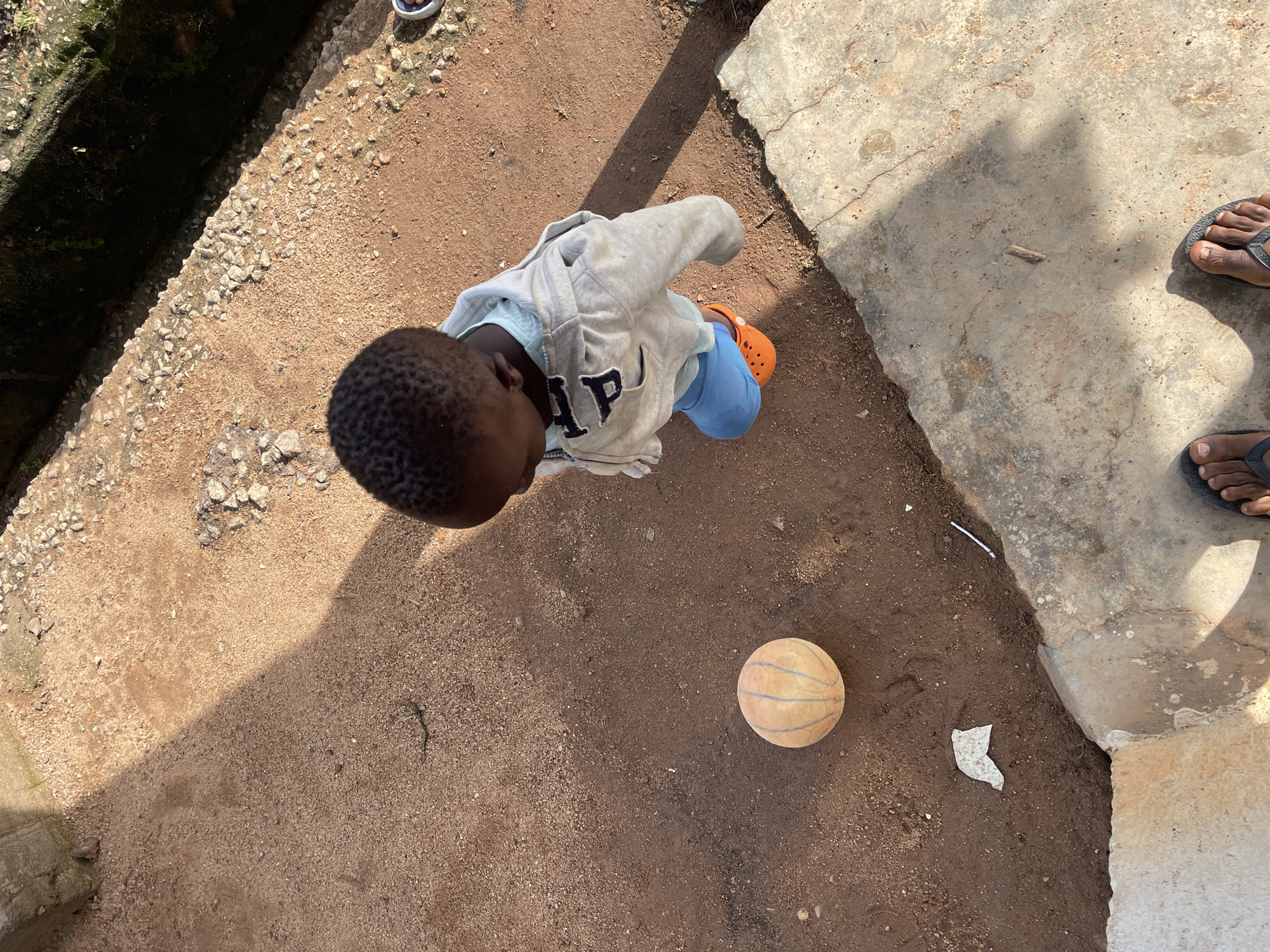 In Barakin Lerawi, Nigeria, a young child with dark, curly hair, wearing a light blue t-shirt and a gray hoodie with "GAP" written on it, walks on a dirt path. The child is also wearing bright orange Crocs with blue insoles. A deflated, light brown and blue striped ball lies on the dirt path to the left of the child. The environment appears to be an outdoor, somewhat unkempt area with loose soil, scattered small rocks, and concrete structures. To the right of the path is a dark, narrow drainage ditch with some vegetation. The ground is a mix of reddish-brown dirt and patches of sand. In the foreground, the feet of an adult wearing dark flip-flops are visible on a concrete surface. A shadow is cast across the scene from an unseen object to the left.