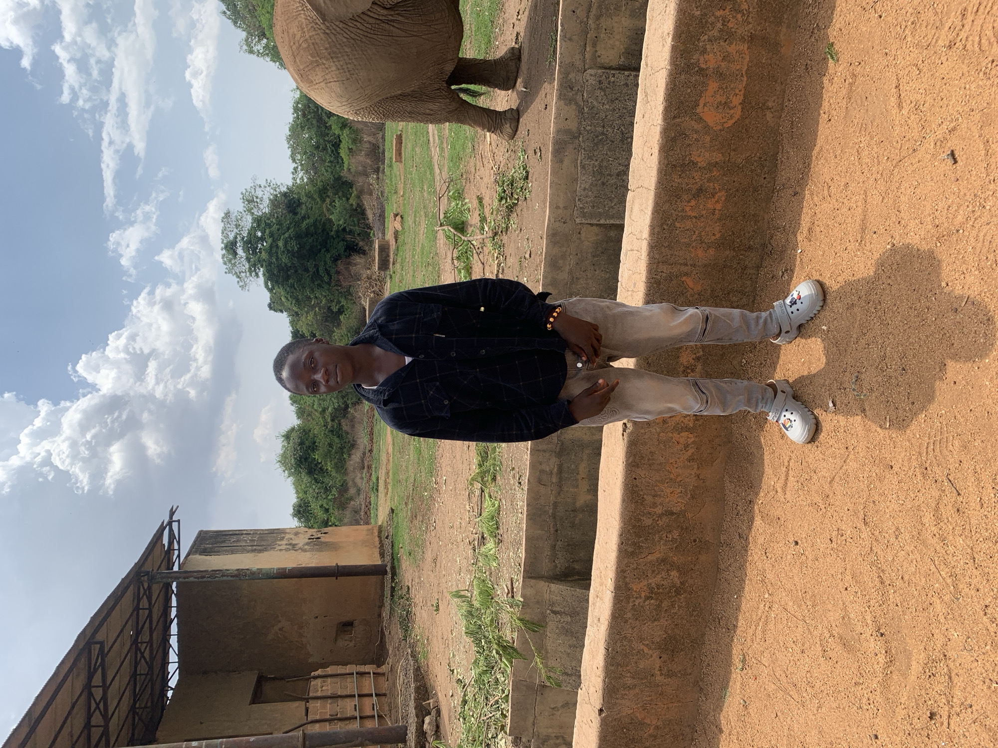 A young man stands beside a low, weathered, concrete wall near a dirt ground. The wall shows signs of age and wear. The man is wearing light-colored jeans, a dark, patterned shirt, and light-colored Crocs with decorative elements. He is positioned on the right side of the frame, casting a shadow onto the dirt.
In the background, an elephant is partially visible, and dense green trees can be seen behind the elephant. To the left, a dilapidated building with a rusted metal roof is visible. The sky is partly cloudy.
The image was taken in Jos, Nigeria.