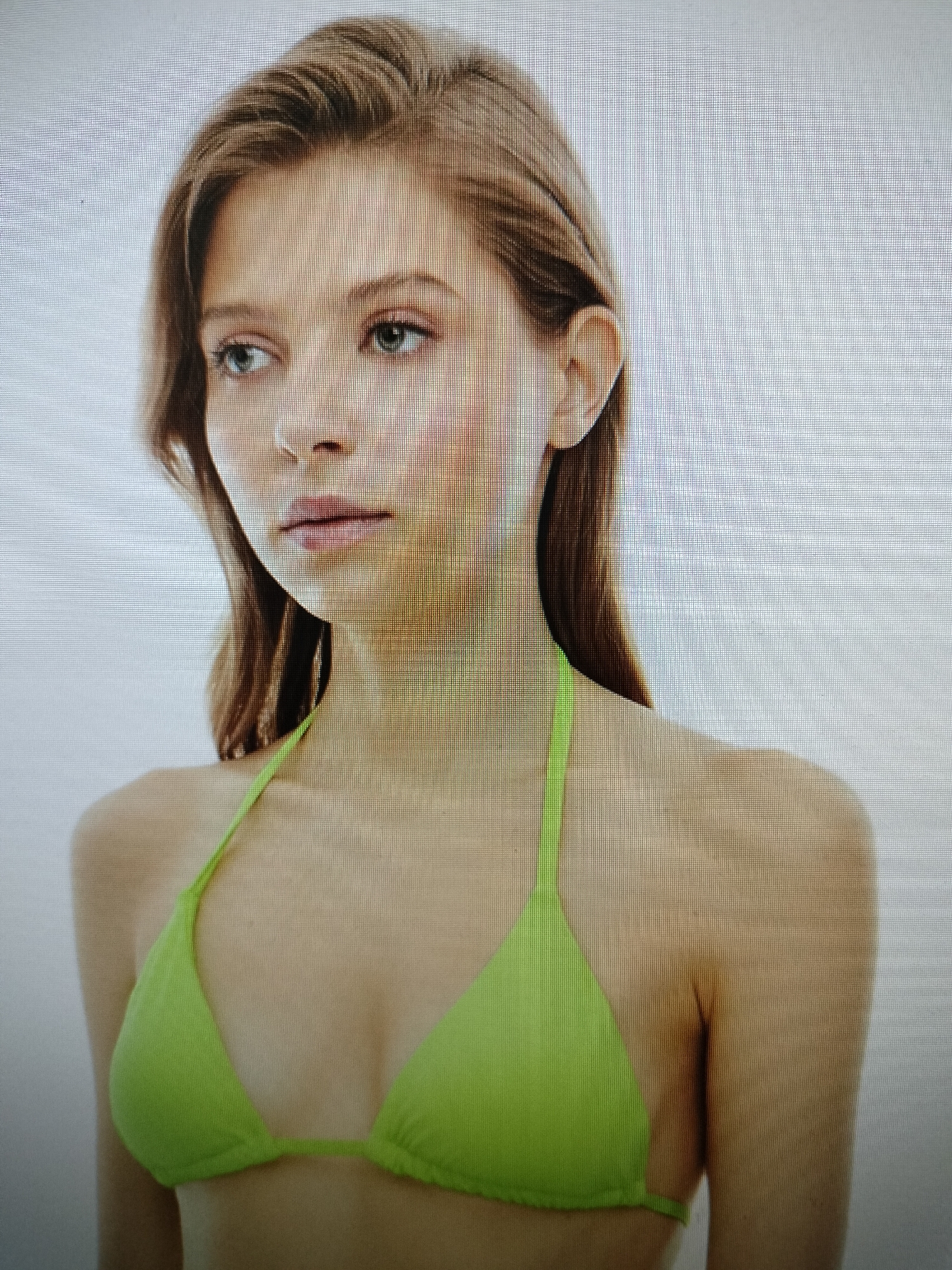 The image provided is a close-up, head-and-shoulders shot of a young Caucasian woman with light brown hair styled in loose waves. She is wearing a bright green bikini top. Her gaze is directed off to the left side of the frame. Her skin tone is fair, and there appears to be some light blush or makeup on her cheeks. The background is a plain, light gray. There is no discernible information within the image that places it in the city of Schaerbeek - Schaarbeek, Belgium.