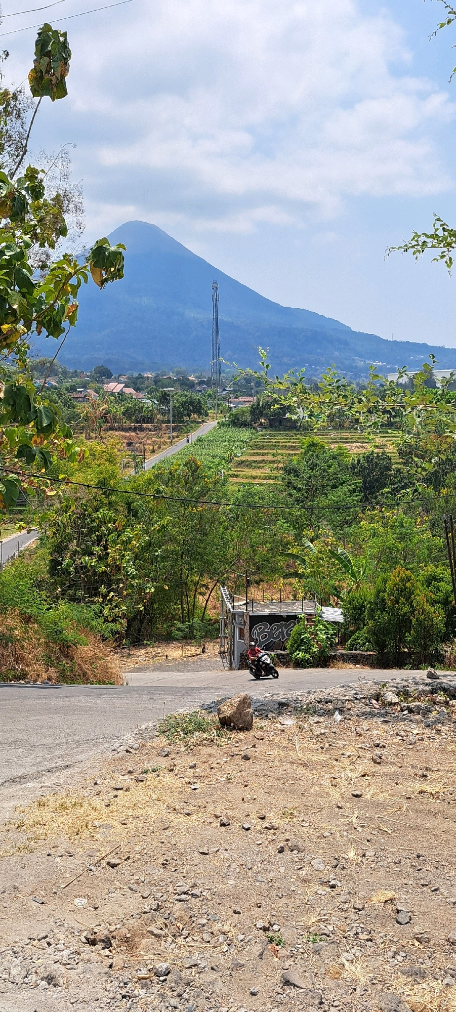 A vertical, eye-level, medium shot captures a rural landscape in Sumber Gedang, Indonesia, with a prominent volcano in the background. The volcano, a muted blue-gray, dominates the upper portion of the frame, its peak slightly obscured by a tall, slender cell tower positioned on its slope. Below the volcano, a village with scattered houses and buildings is visible, nestled amongst lush green vegetation. A road winds its way through the village and towards the foreground. Terraced fields, displaying a mix of green and golden hues, occupy the middle ground, suggesting agricultural activity.
In the foreground, a person on a black motorcycle, wearing a red shirt, is riding on the road. The motorcycle is turning towards the right side of the frame, navigating a slight incline. To the right of the road, a small, utilitarian structure with a graffiti-covered wall is visible. The ground in the immediate foreground is dry and uneven, covered with gravel, rocks, and sparse, dried grass. Trees with green leaves frame the scene from the left and right sides, with some foliage in the foreground partially obscuring the view. The sky is bright blue with scattered white clouds. The overall impression is one of a peaceful, rural setting with a blend of natural beauty and human infrastructure.