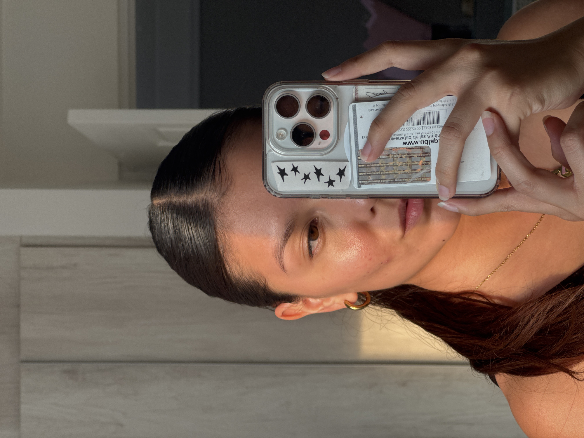 A woman is holding a smartphone in front of her face to take a selfie. The phone has a clear case with black star stickers and what appears to be a credit card or license slipped into the back. The woman has dark hair pulled back, a gold hoop earring in her left ear, and a delicate gold necklace. Her skin appears to have a natural glow. The background consists of what may be a wall, part of a cabinet, and a doorway. The environment is generally bright with natural light.
