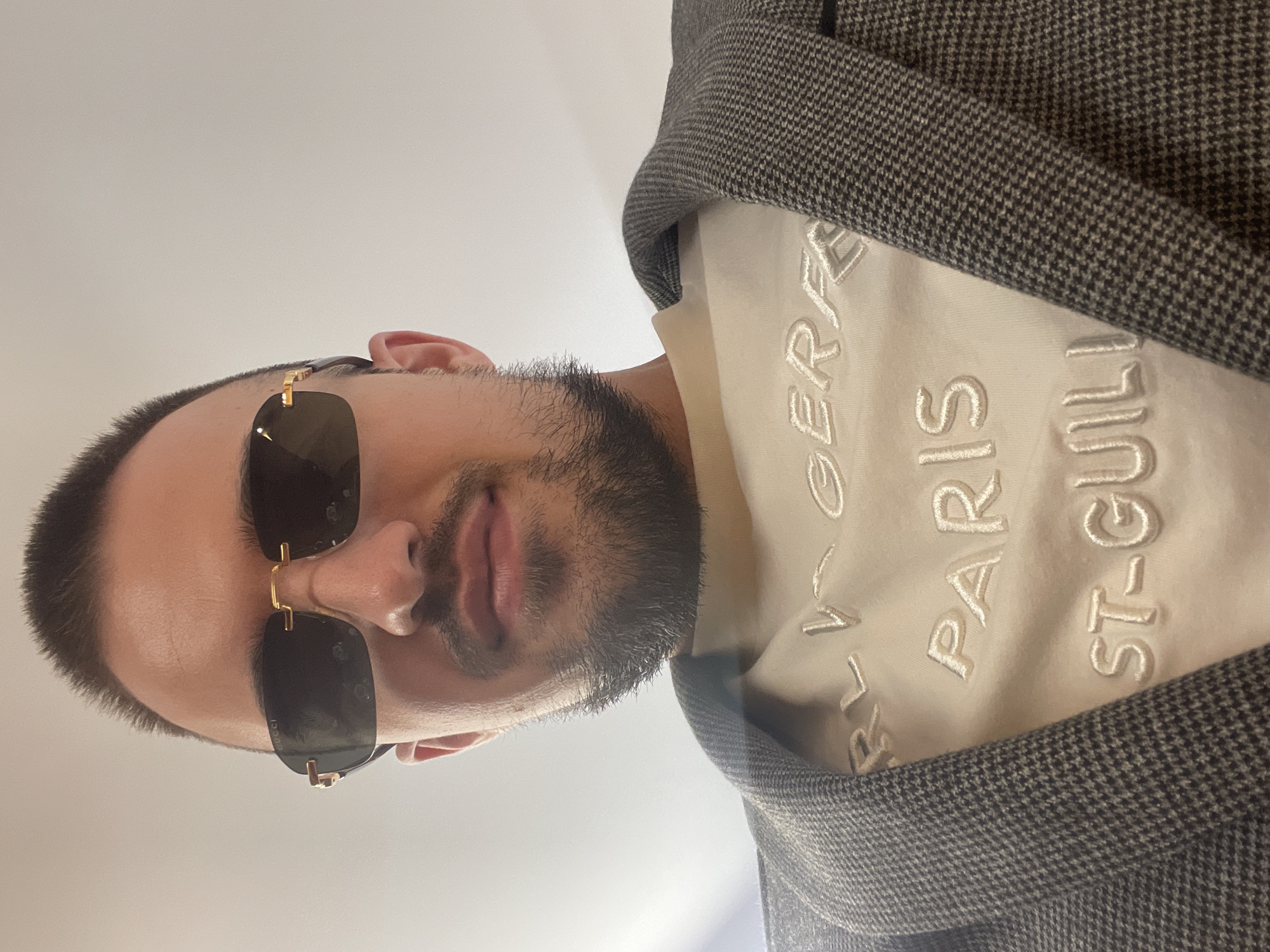 A man is depicted in a close-up, slightly angled, selfie-style shot. He is wearing dark sunglasses with gold-colored frames, a white shirt with raised lettering that includes the words "PARIS" and "ST-GUILL", and a grey herringbone-patterned jacket. The location is not discernible from the image.
