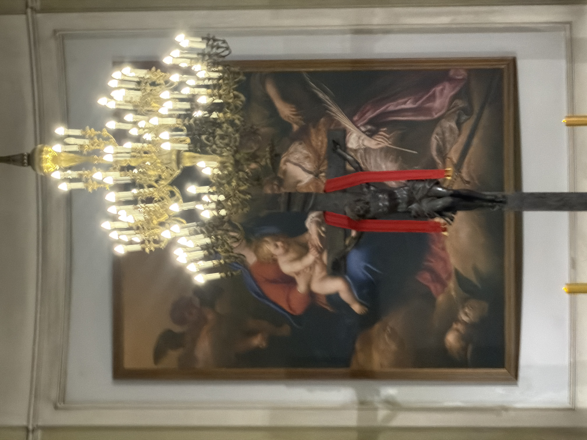 Captured from below, the image portrays an interior scene in Saint Petersburg, Russia. A large, ornately decorated chandelier with multiple lit candles dominates the left foreground. Behind the chandelier, mounted on the wall, is a framed painting. The artwork depicts figures including a woman holding a baby and winged figures, rendered in a classical style with muted colors. A dark crucifix, draped with a red cloth, horizontally bisects the center of the frame, partially obscuring the painting. The background consists of a neutral-toned wall with decorative moldings.