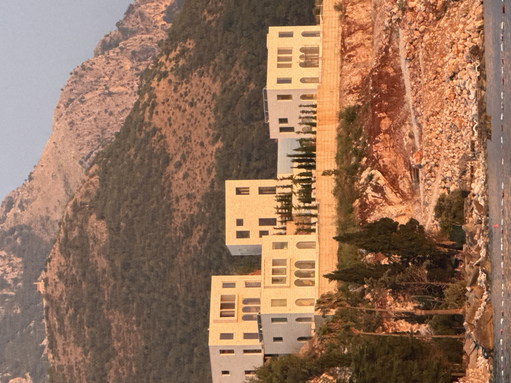 The image depicts a coastal scene in Kamenovo, Montenegro, with a cluster of modern, light-colored buildings set against a backdrop of a rugged, tree-covered mountain. The buildings feature clean lines, flat roofs, and large windows, some with arched architectural elements. They appear to be multi-story structures, possibly a resort or residential complex.
In the foreground, the rocky shoreline meets the water. The water surface shows a pattern of white and red buoys or markers, indicating a designated swimming area or a boundary. Scattered rocks and pebbles are visible along the coastline and on the embankment leading up to the buildings.
Vegetation is present in the midground, including several tall, slender cypress trees and some palm trees, interspersed with lower-lying shrubs and greenery. The mountain slopes behind the buildings are covered with dense, dark green vegetation, transitioning to more sparse, reddish-brown scrub and exposed rock faces higher up. A small, isolated structure, possibly a chapel or a small building, is visible on a prominent ridge near the summit of the mountain.
The lighting suggests it is either early morning or late afternoon, with warm, golden light illuminating the scene and casting soft shadows. The sky is a pale, washed-out blue, indicating a clear day. The overall impression is one of a serene, upscale coastal development nestled within a natural, mountainous landscape.