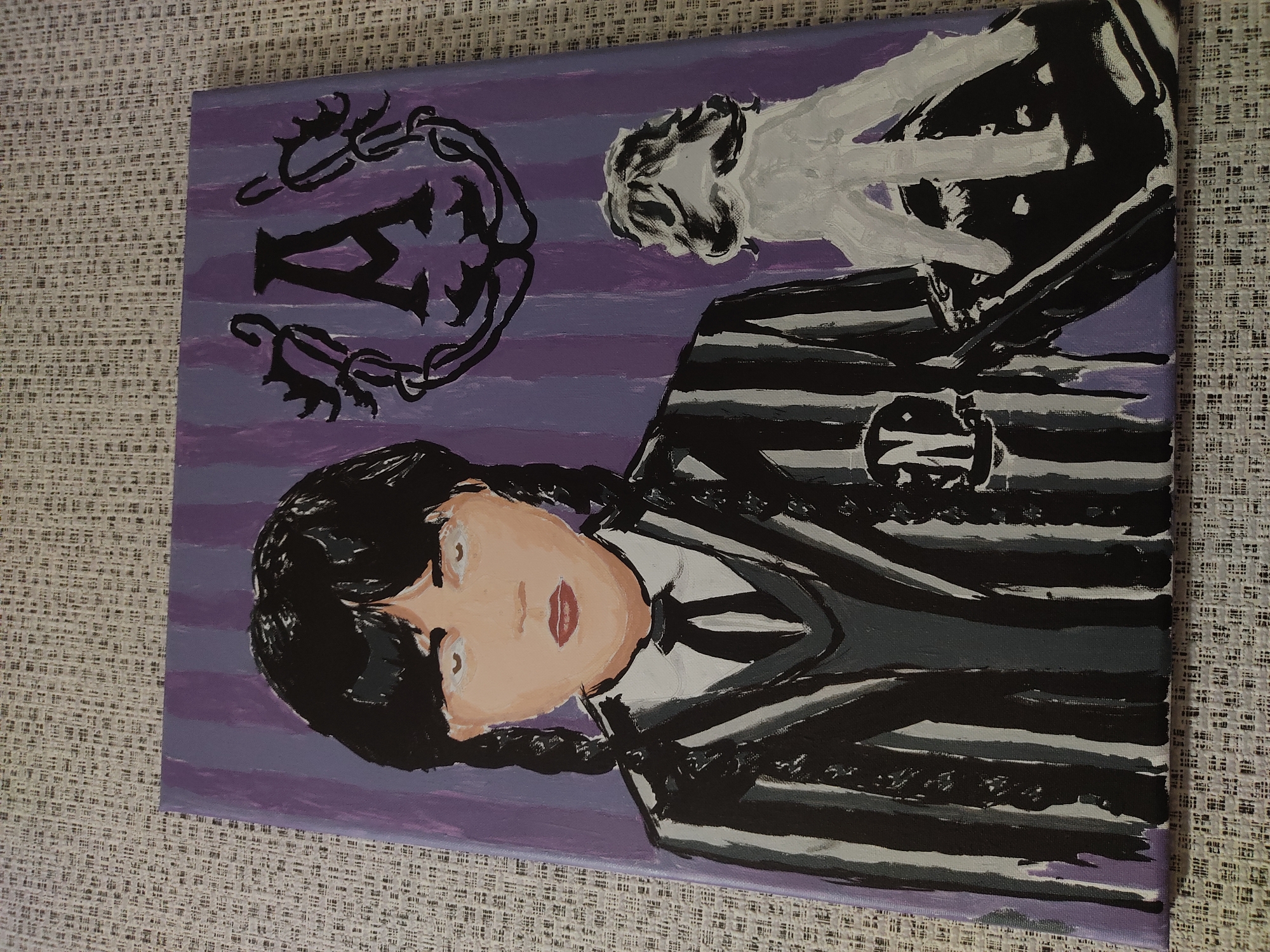 The image shows a painting of Wednesday Addams on a canvas with a purple striped background. She is depicted with dark braided hair, pale skin, and white eyes. She is wearing her signature black and white striped uniform with a tie and a gray V-neck sweater. On her left breast is a circular emblem with the letter "N." To her right, a white, disembodied hand is prominently featured. In the upper right corner of the painting, a black emblem resembling a bat or crest with the letter "A" in the center is visible. The painting is placed on a textured, light-colored surface. There is no information available about the city of Horbanivka, Ukraine in relation to this image.