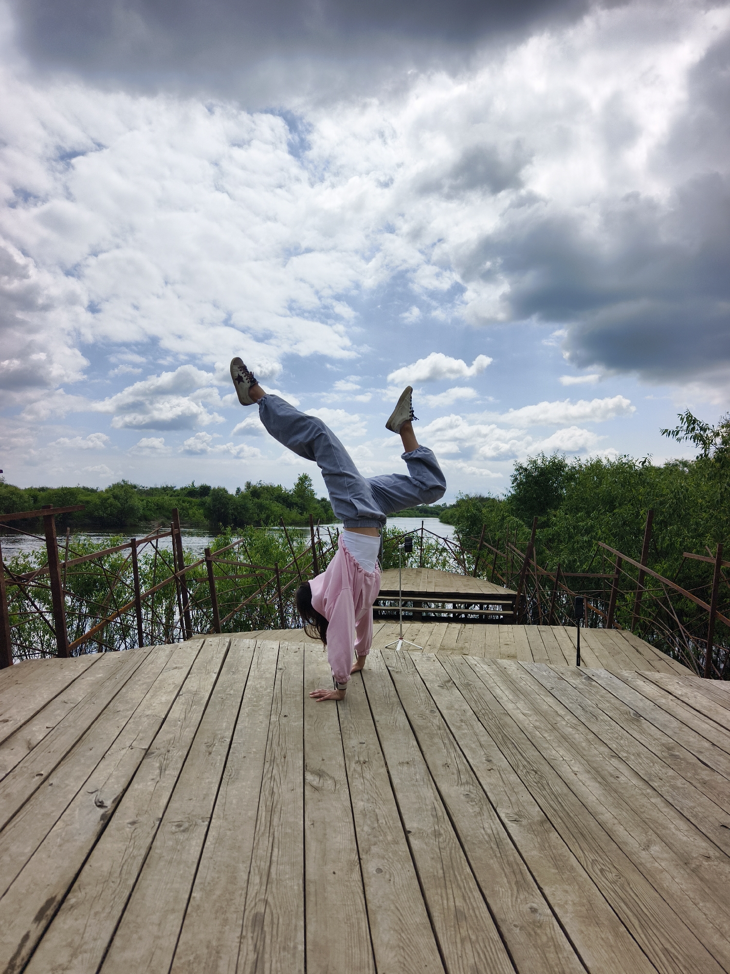 A person performs a handstand with legs raised in a V-shape on a wooden deck overlooking a river. The person is wearing a pink hoodie, grey pants, and white underwear. They are supported by their hands with their body inverted. The wooden deck is made of weathered planks. Beyond the deck, a rusty metal fence runs along the edge of the river, with lush green trees and foliage lining the opposite bank. The river is calm and reflects the sky, which is filled with dramatic, swirling clouds. The location is the city of OnoKhino, Russia.