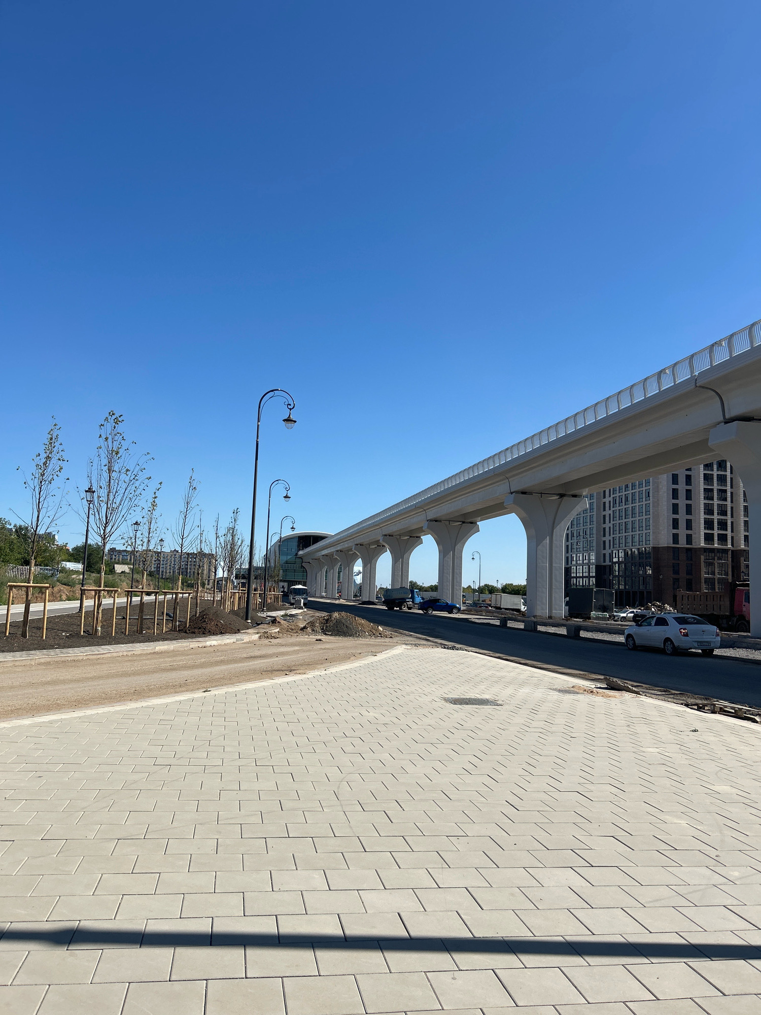 The image depicts an outdoor scene in Astana, Kazakhstan, under a clear blue sky. In the foreground, a newly paved area with light gray rectangular pavers is visible, with a shadow cast across it. Behind the paved area, a dirt road or construction site is present, bordered by young trees with wooden supports. Several ornate streetlights are positioned along the road. Further in the background, a modern elevated structure, likely a monorail or elevated highway, supported by massive concrete pillars, dominates the scene. Adjacent to this structure is a tall, modern building with numerous windows. Several vehicles, including a white car and a blue car, are on the road. The overall impression is one of ongoing urban development and infrastructure construction in a city setting.