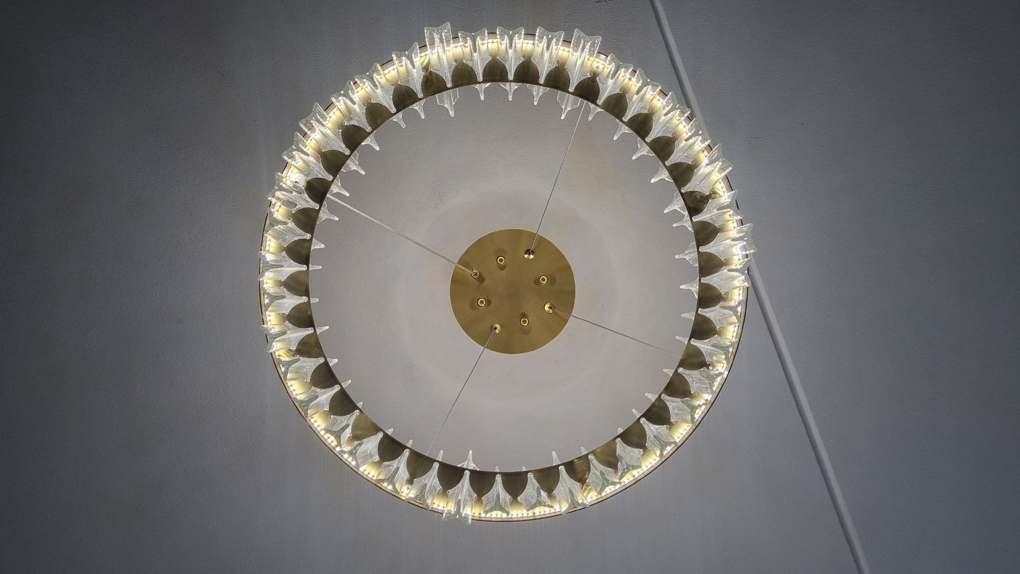 A close-up, worm's-eye view shows a circular chandelier hanging from a light gray ceiling. The chandelier has a gold-toned metal frame and is adorned with numerous clear, pointed glass elements that are lit from within, giving off a warm, yellow light. The chandelier is suspended by thin wires connected to a central gold-toned circular canopy attached to the ceiling. The ceiling itself has a subtle texture and a faint diagonal line across its surface. No people or other objects are visible in the scene. The location is specified as Верзилово, Russia.