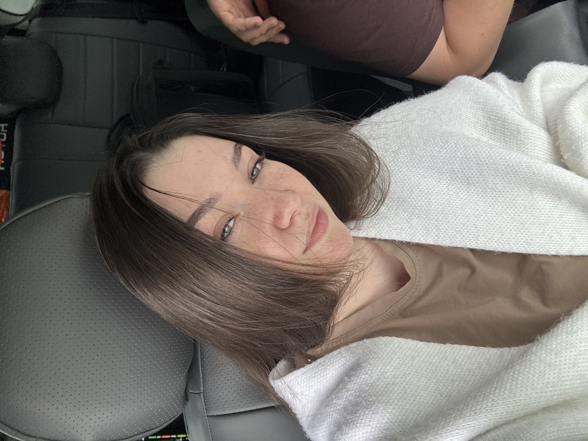 The image shows a young woman with brown hair and green eyes, wearing a beige t-shirt and a white cardigan. She is sitting in the back seat of a car, with dark grey leather seats visible. The car appears to be parked, and the lighting suggests it's daytime. Her expression is relaxed. The image does not provide enough information to determine the specific location within the city of Perm, Russia.