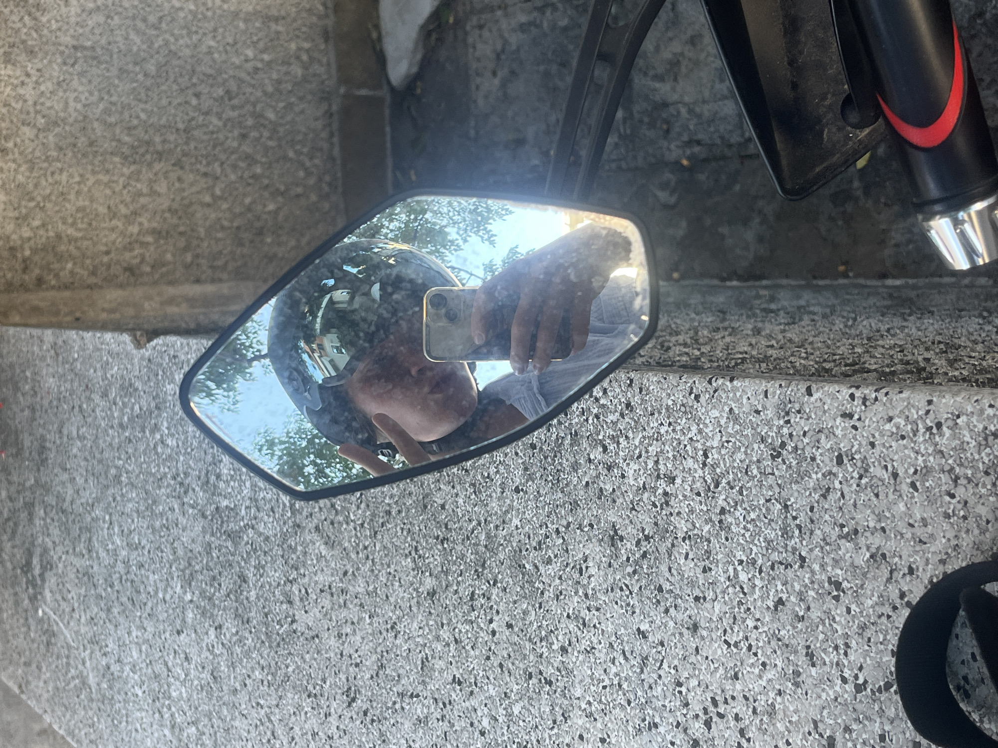 The image shows a motorcycle mirror reflecting a person wearing a helmet and holding a phone to take a selfie. The mirror is attached to a motorcycle part with a red and black accent. The motorcycle is parked on a gray, textured surface. The background is obscured by the reflection in the mirror.
The motorcycle appears to be located on/near a concrete curb or step. The location in the city of Budva is unknown.