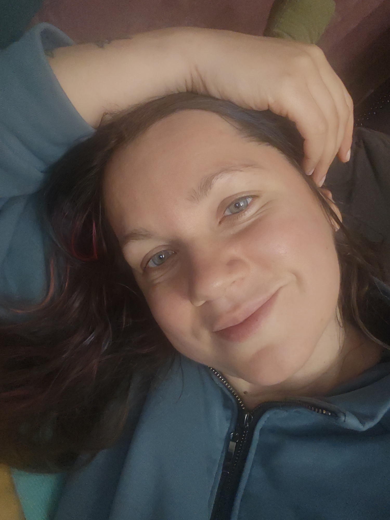 A young woman with light skin and blue eyes is lying down, looking directly at the camera with a slight smile. Her hand is resting on her forehead. She has dark brown hair with a hint of pink. She is wearing a teal zip-up jacket. The background is out of focus. No information suggests a location in Свистуха (кузяевский С/о), Russia.
