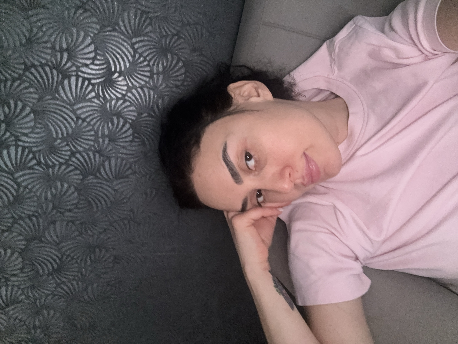 The image captures a person, likely female, lying down indoors on a gray surface, possibly a couch or bed. The individual is positioned diagonally, with their head towards the upper left of the frame and their body extending towards the lower right. They have dark, possibly curly hair, prominent dark eyebrows, and a light complexion, looking directly at the camera with a calm and slightly smiling expression.
The person is wearing a light pink, short-sleeved t-shirt. Their left hand is gently resting near their chin, and a small, dark tattoo, appearing to be a floral or abstract design, is visible on their left forearm.
To the left of the person, a dark gray wall or headboard is visible, featuring a distinct textured pattern composed of numerous swirling, fan-like motifs that create a sense of depth. The background on the right side of the image consists of the plain gray of the seating surface. The lighting is soft and even, suggesting a comfortable indoor environment. There are no discernible visual cues within the image itself to indicate the provided location context of Tbilisi, Georgia. No visible text is present in the image.
