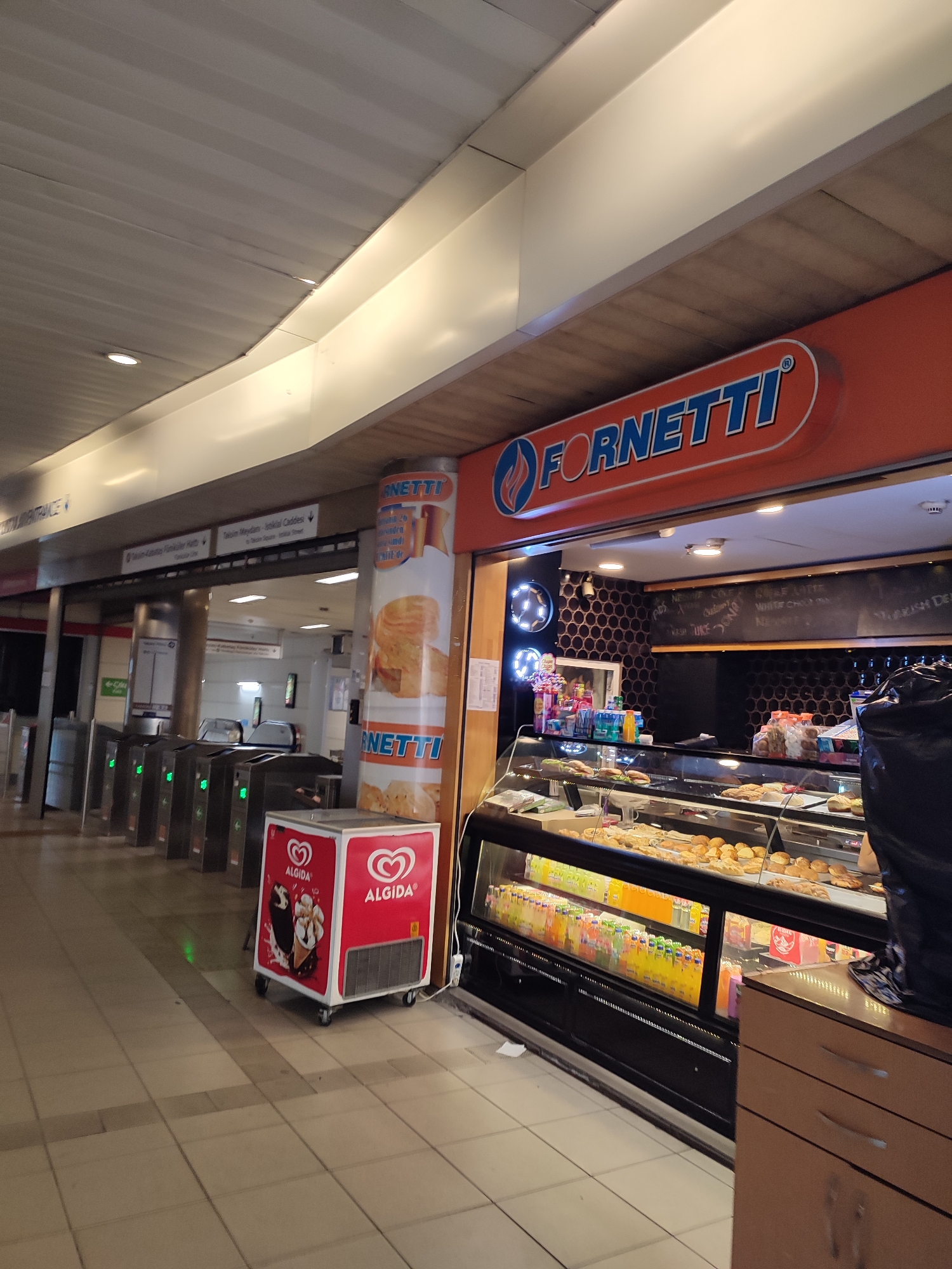The image shows a Fornetti bakery and cafe located in the city of Beyoğlu, Turkey. In the foreground, there is a red and white ice cream freezer with "Algida" branding. Behind the freezer, there are ticket barriers that appear to be in a subway or train station. To the right of the ticket barriers and ice cream freezer is the Fornetti shop. The shop features a prominent orange sign with the "FORNETTI" logo in blue lettering. Inside the shop, a display case is filled with baked goods and bottles of colorful beverages. A blackboard behind the counter displays a menu. The overall environment suggests a public transit hub with food service available.