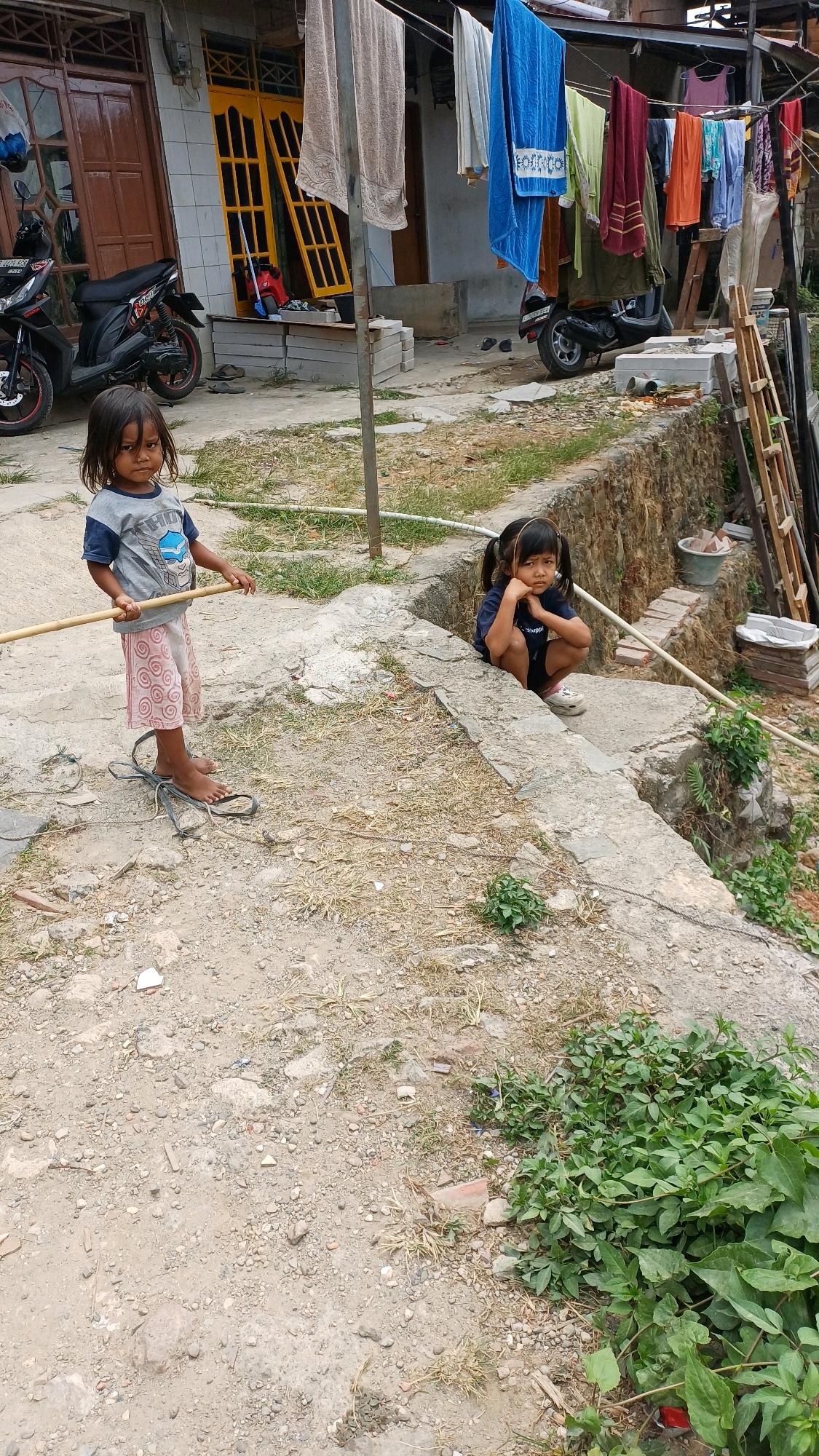 In Citeureup, Indonesia, two young girls are pictured outdoors in a residential area with a dirt and gravel ground. The girl on the left, wearing a grey t-shirt with a blue graphic and pink patterned shorts, stands holding a wooden stick. She has dark hair and is barefoot. The girl on the right, in a dark blue t-shirt and black shorts, is crouched down, resting her chin on her hands. She also has dark hair styled in pigtails. Behind them, laundry is drying on a clothesline strung between poles. Various buildings, a parked motorcycle, and construction materials are visible in the background. The environment appears to be a humble neighborhood.