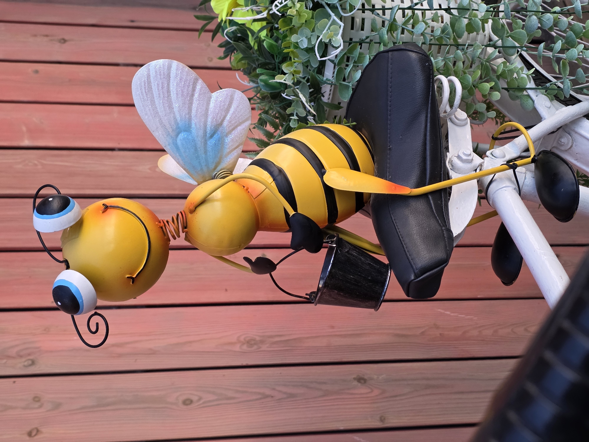 An ornamental, yellow, cartoonish bee made of metal is perched on the black seat of a white bicycle. The bee has large, cartoonish eyes with blue and black pupils, and it is holding a small, black bucket in its left hand. Its wings are iridescent and light blue, and its body is striped with black and yellow. The bicycle has white handlebars and a white frame. The bee is positioned in front of a reddish-pink wooden fence. To the right of the bee, there is a white planter filled with lush green foliage. The scene is outdoors and appears to be in a garden or outdoor decorative setting. The specific location in the city of Chaikovsky, Russia, is not discernible from the image itself.