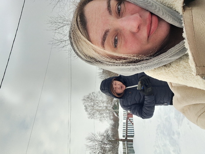The image depicts two individuals outdoors in a snowy, rural setting in Полянки, Russia, under an overcast sky. A blonde female subject, positioned in the right foreground, wears a cream-colored, shearling-lined coat and a light grey headscarf, looking directly at the camera with a neutral expression and slightly reddened cheeks. Behind her, to the left, a male subject is observed wearing a dark blue hooded puffer jacket and black gloves, focusing on a smartphone held in both hands; his mouth is partially open. The ground is uniformly covered in undisturbed snow. In the mid-ground, a painted wooden picket fence (white and blue) partially separates the subjects from a sparse winter landscape featuring bare deciduous trees and a distant building. Overhead, two utility lines are visible against the grey sky.