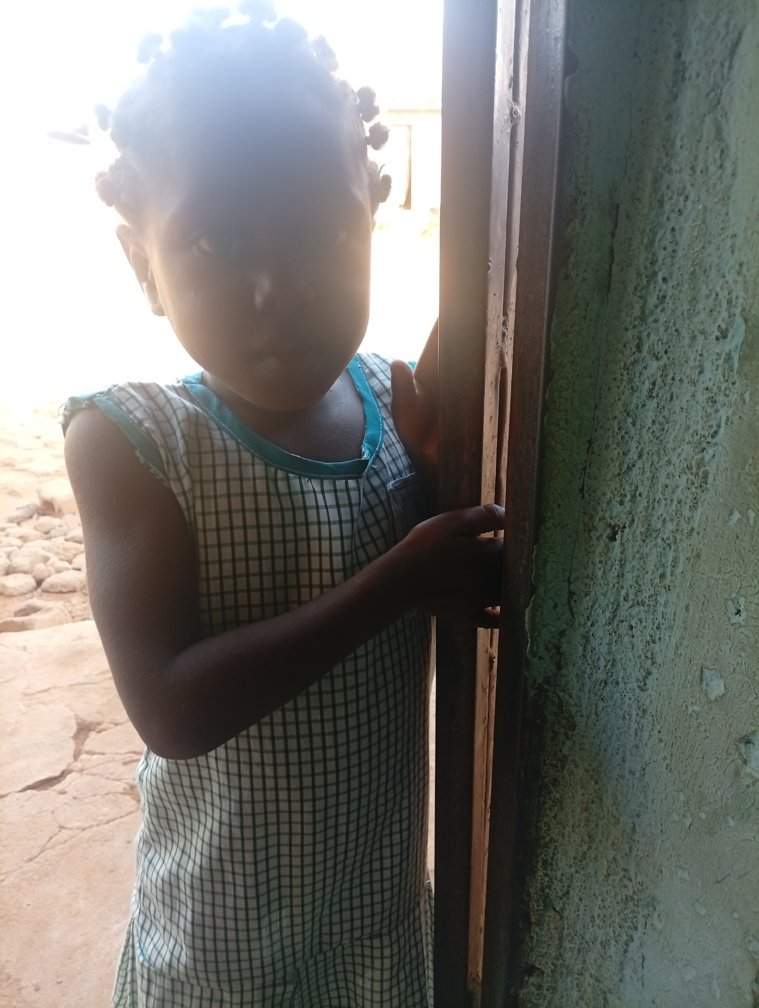 A young child is standing partially behind a door. The child has dark skin and a hairstyle of small, tightly wound braids or knots. They are wearing a sleeveless dress or top with a checkered pattern and a light blue trim around the neck and arms. The child's expression is difficult to discern due to the lighting and the angle. The door is made of dark-colored wood or metal, and the wall next to it has a rough, textured surface with layers of peeling paint, appearing in shades of green and white. The background is out of focus but seems to show an outdoor area with a cracked, possibly concrete or earthen surface. The location is unknown, and the city is also unknown.