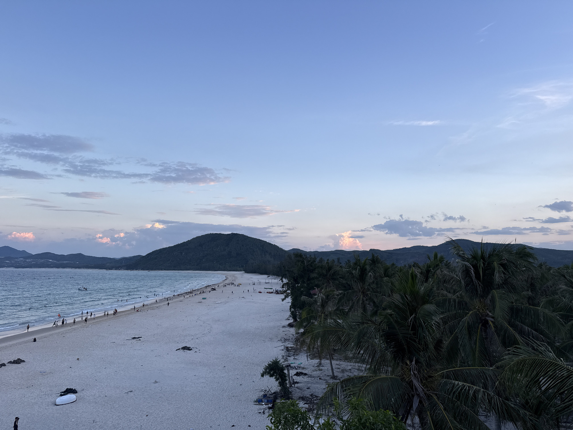 A high-angle, wide shot captures a beach at dusk. The sky is a gradient of pale blue to a soft pink, with scattered clouds. The ocean, a deep blue, gently laps against a wide, sandy beach. Numerous people are dotted along the shore, some walking, others wading in the water. A few boats are visible in the distance. The beach is lined with lush green palm trees, their fronds obscuring a portion of the background. In the far distance, dark, tree-covered mountains form a silhouette against the twilight sky. The sand is a light, powdery white, with a few scattered rocks and debris. One white object, possibly a surfboard or kayak, lies on the sand.
