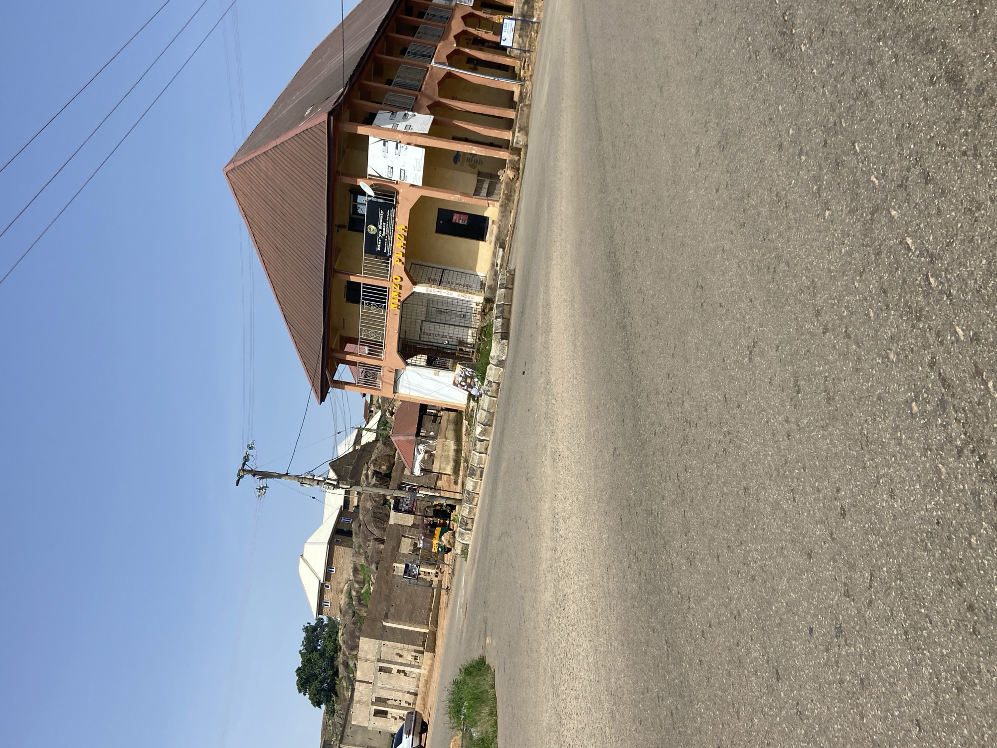 A daytime, eye-level shot captures a street scene in Jos, Nigeria. In the foreground, a paved road with patches of green grass on the left curves into the distance. Dominating the mid-ground is a two-story building with a brown, sloped roof and an orange-colored exterior. The building features a balcony on the second floor, supported by arched pillars on the first floor, which also has glass-paneled shopfronts. Signage on the building includes "NINZO PLAZA" in yellow letters and a smaller sign for "Mary's Beauty Institute." Adjacent to this building on the left is an unfinished brick structure with exposed rebar. Further in the background, a cluster of houses with white roofs are situated on rocky terrain, a large green tree standing to the left. A utility pole with wires is visible on the right side of the frame. Several vehicles, including a white car and a yellow and green auto-rickshaw, are present on the street. The sky is clear and blue.