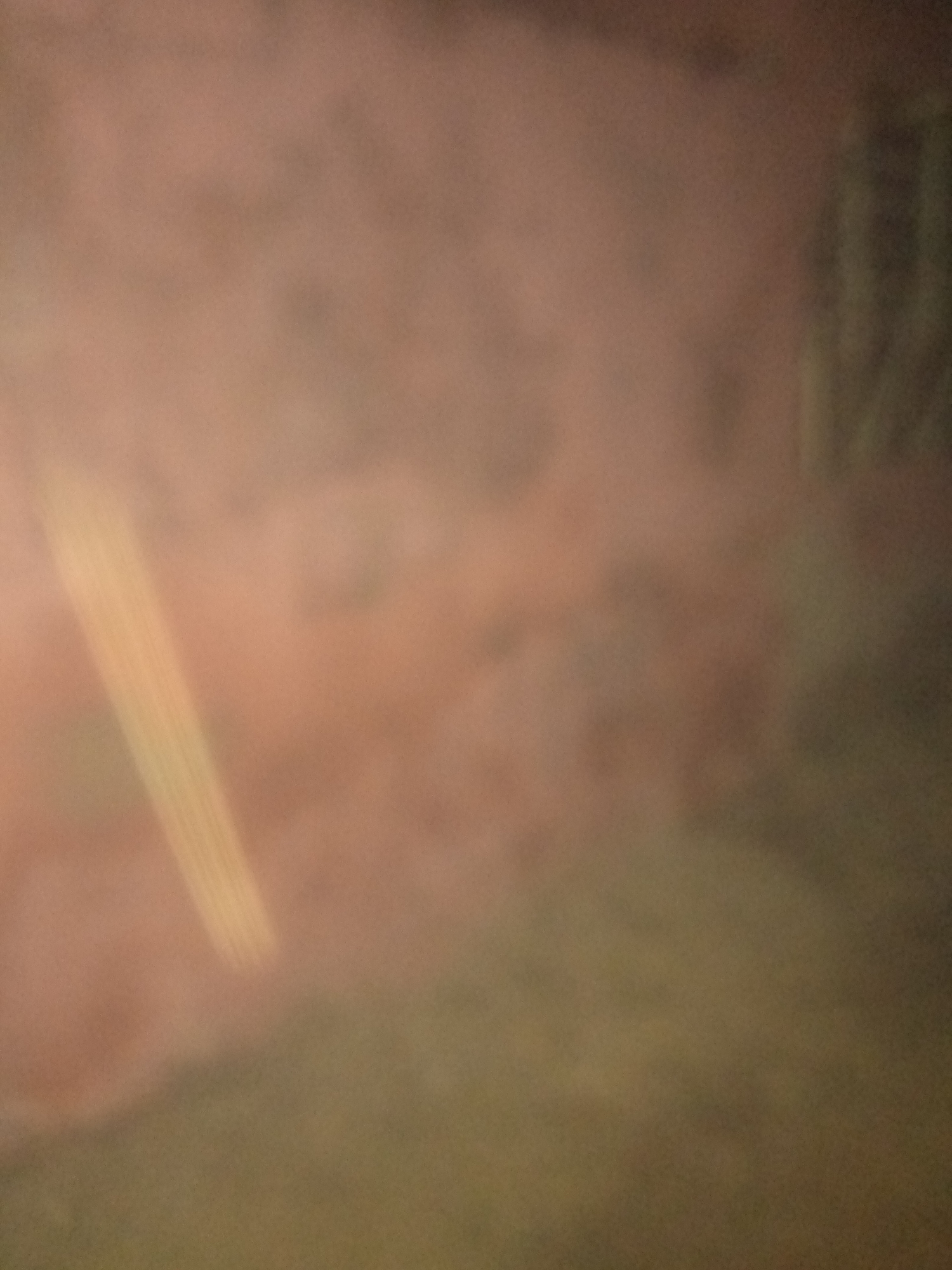 The image displays a very out-of-focus, hazy, abstract scene with a predominantly pinkish-brown hue. In the left portion of the image, a vertical, yellowish object with faint parallel lines is visible, partially obscured by the haze. The background is a muted blend of pinkish-brown and darker, muted green tones. No discernible location, people, or specific objects can be identified with certainty due to the extreme blurriness.