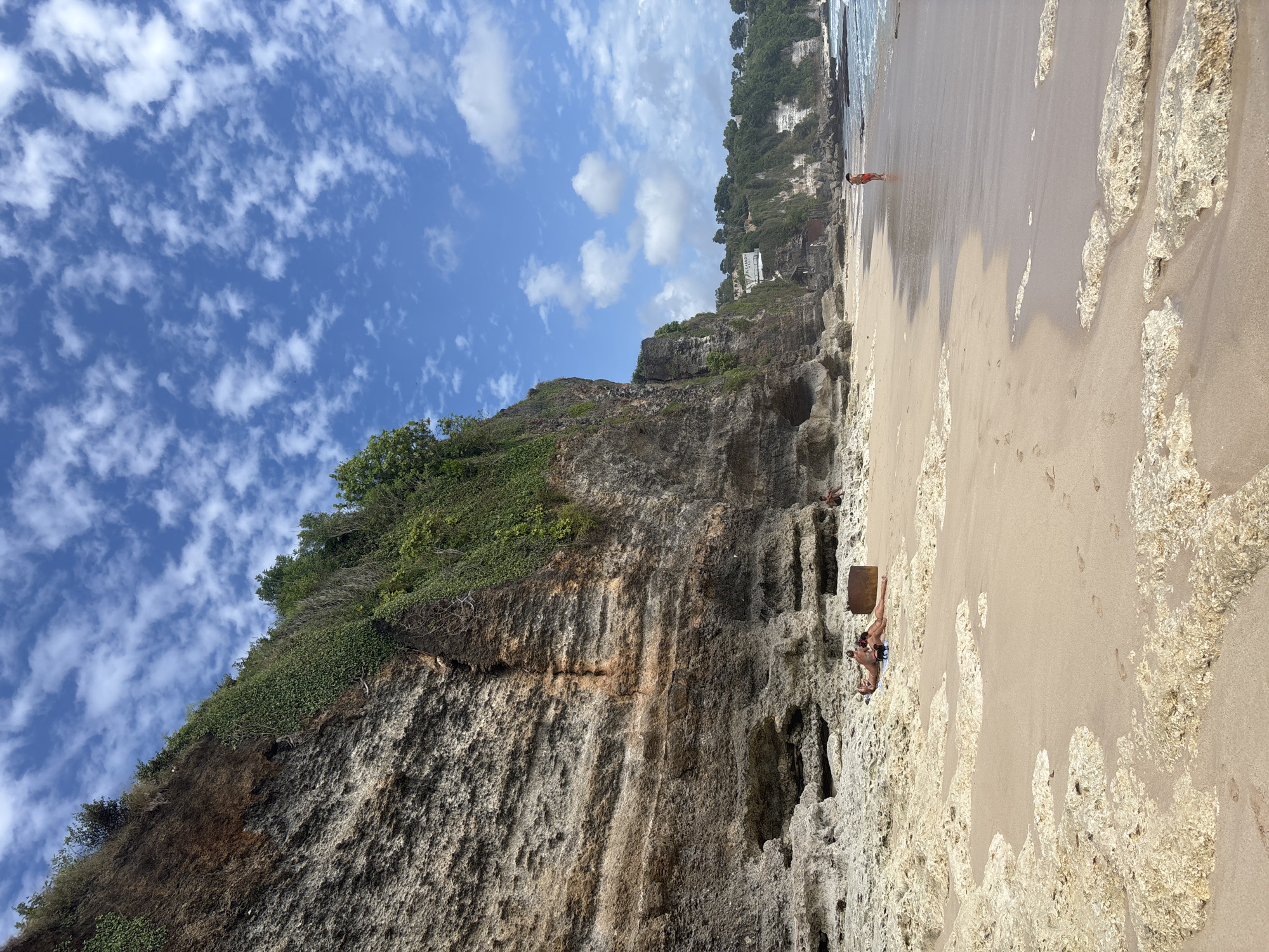 In Kuta Selatan, Indonesia, a serene beach scene unfolds with a dramatic cliff face dominating the left side of the frame. The cliff is rugged, layered with various shades of brown and tan rock, and sparsely vegetated with green foliage clinging to its upper reaches. At the base of the cliff, a sandy beach is dotted with light-colored rocks and pebbles. In the foreground, the sand is wet from the receding tide, with footprints visible.
On the beach, two individuals are lying down, appearing to sunbathe. They are dressed in swimwear. A dark, cylindrical object, possibly a rusted barrel, is situated near them. Further down the beach, a single person in red shorts stands near the water's edge. In the distance, the coastline curves, with more green-covered cliffs and a few buildings visible atop them. The sky above is a vibrant blue, streaked with wispy white clouds. The ocean meets the shore with gentle waves.