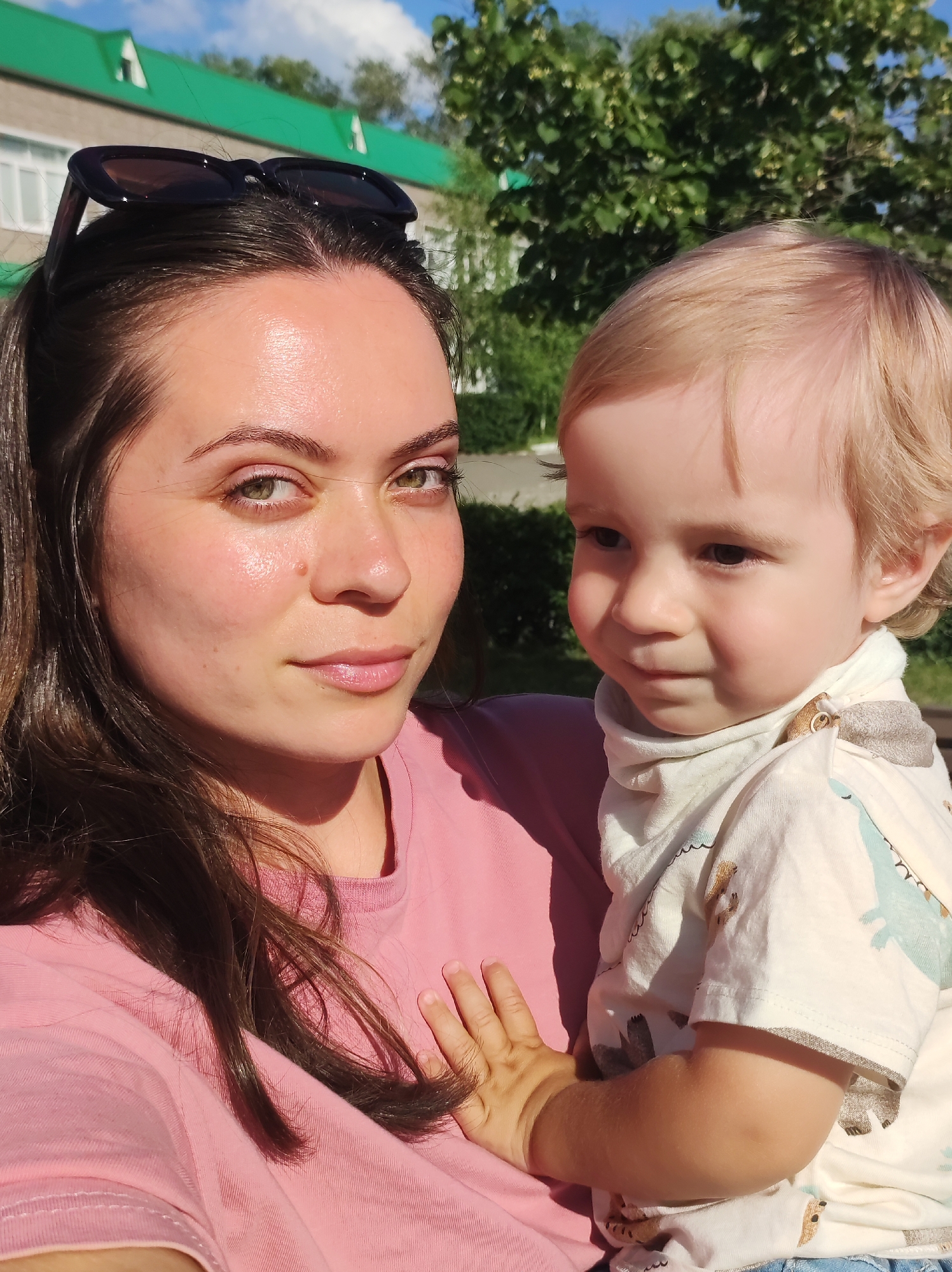 This is a medium shot, taken outdoors in daylight, of a woman holding a young child. The woman, positioned on the left, has long, dark brown hair pulled back, and is wearing a pink t-shirt. She is looking directly at the camera with a neutral expression. Sunglasses are perched on top of her head. The child, on the right, has light blond hair and is wearing a white t-shirt with a light blue and grey dinosaur print and a white scarf. The child is looking to their left, away from the camera, with a slightly wistful expression. The background is slightly blurred, showing a building with a green roof and windows, and lush green trees under a blue sky with white clouds. The overall lighting suggests a sunny day. The location is identified as Chaikovsky, Russia.