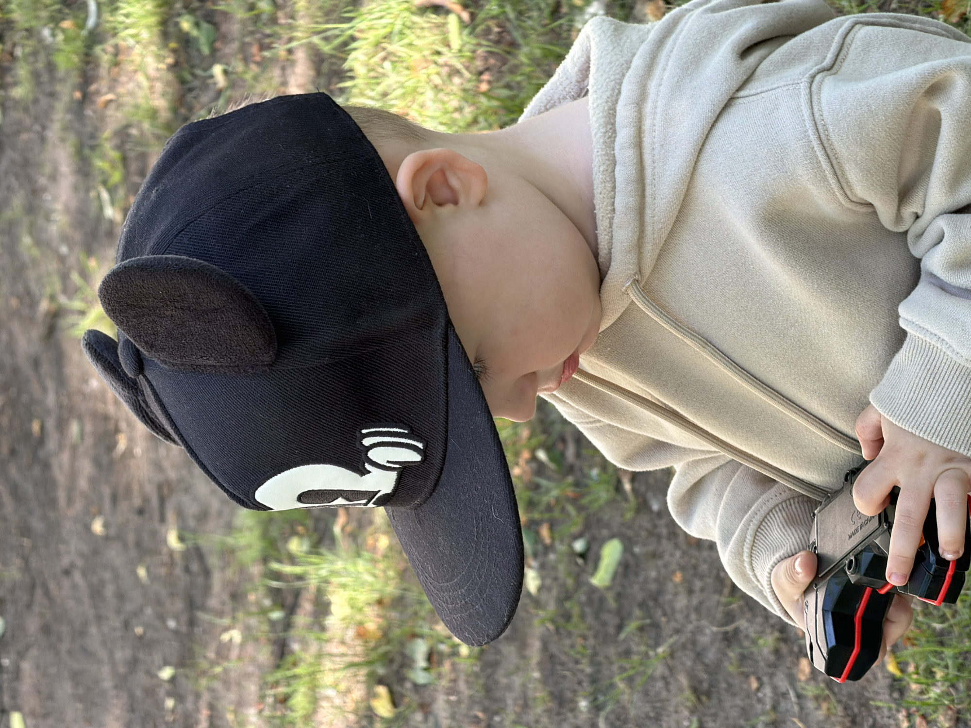 A young child, presumably in the city of Chaikovsky, Russia, is depicted outdoors, observed from a slightly elevated perspective. The child wears a black baseball cap featuring attached dark brown, fuzzy ear-like appliques and a partially visible white emblem on the front, appearing to be a stylized letter "R" or "B" with an accompanying graphic detail. The child's head is tilted downward, obscuring the full face, while a portion of the chin and lips are visible. A light beige or cream-colored hooded sweatshirt with visible drawstrings covers the child's torso and arms. Both hands hold a black and red electronic device, resembling a toy or controller, with "MADE IN CHINA" faintly imprinted on its rear surface. The child's focus is directed entirely toward this object. The ground beneath consists of loose dirt, interspersed with patches of green grass and scattered dry leaves.