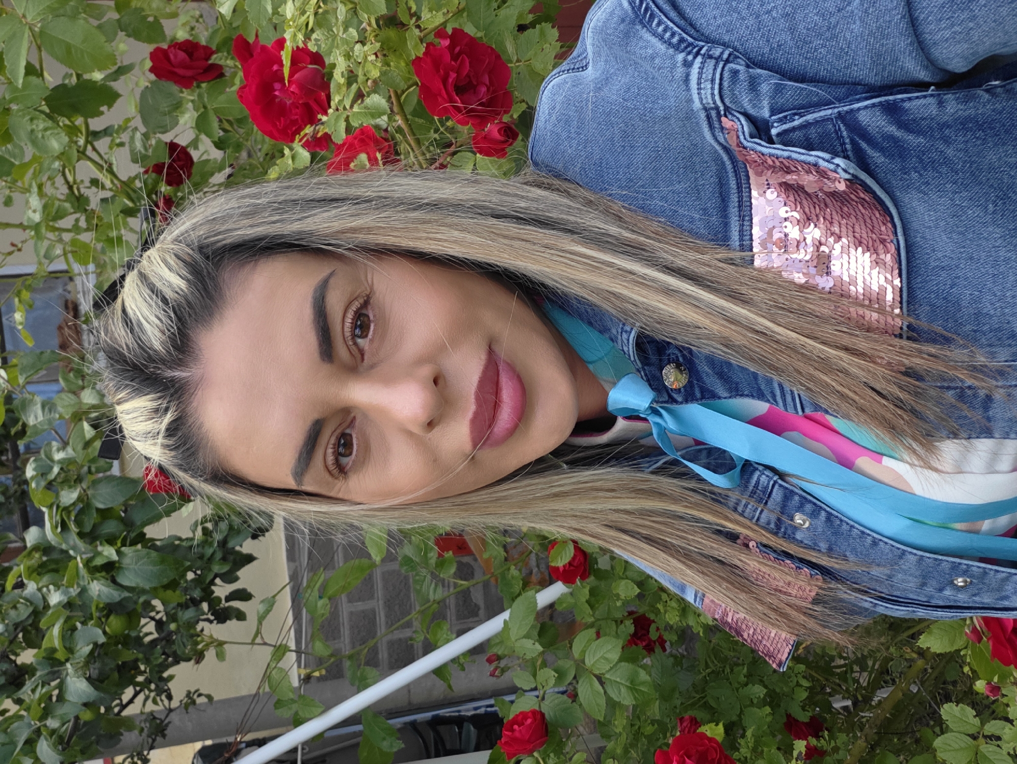 The image is a medium shot portrait of a blonde woman in Ravnogor, Bulgaria, standing in front of a rose bush with red roses. She has her hair pulled up and parted in the middle. She is wearing a denim jacket with pink sequin accents on the pockets and a colorful shirt underneath with a blue scarf tied around her neck. The background shows a house with a brick wall and greenery. The lighting suggests it is daytime.
