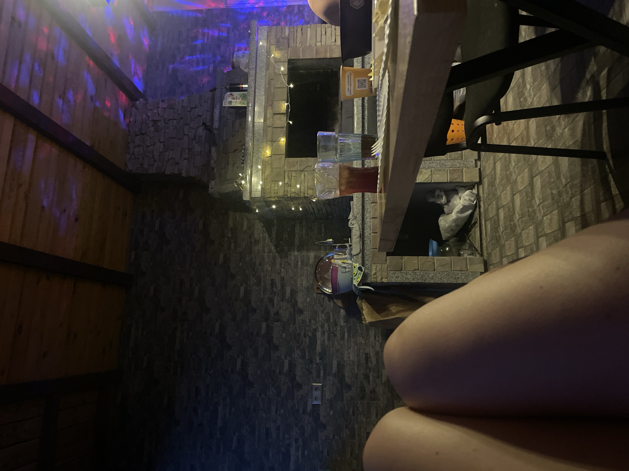 The image captures a dimly lit indoor or semi-enclosed outdoor area in Petrivka, Ukraine, from a low perspective, likely of a person seated at a table.
In the foreground, the bare upper thighs of a person occupy the lower left and central portions of the frame. A wooden table extends into the scene from the lower right. On the table, from left to right, are two tall glasses, one containing a reddish-brown liquid with ice, and another containing a darker liquid. A yellow disposable cup with Ukrainian text reading "ЇЖА З ДОСТАВКОЮ ЧЕРКАСИ" (Food with delivery Cherkasy) is visible, along with a black bowl and a white scalloped-edge plate or napkin with a fork resting on it.
The midground features a prominent stone-tiled wall on the left, equipped with a double electrical outlet. A substantial built-in structure, appearing as a fireplace or grill unit, dominates the central area. It is constructed from light-colored stone and features a hearth. A string of warm white decorative lights is draped across its mantelpiece. Resting on the mantelpiece are a small potted item, a dark bottle, and a glass jar. To the immediate left of this structure, a small countertop with a sink, a large clear bowl, a smaller purple bowl, and a white cloth adorned with an avocado pattern are visible. Beneath the table, a black metal chair stands, with an orange plastic basket positioned beneath it. A bundle of white material rests on the stone floor to the left of the chair.
The background reveals a wooden plank ceiling supported by dark wooden beams. The ceiling and upper right wall are illuminated by dynamic red, blue, and purple colored lights, casting reflective patterns across the surfaces. The walls throughout the space are faced with dark grey or brown rectangular stone tiles.