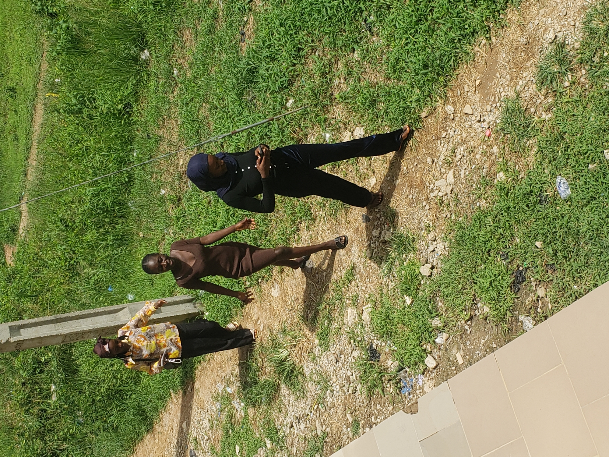 In the city of Gumel, Nigeria, three women are captured from a high angle walking on a dirt path beside lush green vegetation and a concrete pole. The woman in the foreground, on the right, wears a dark blue hijab and a black long-sleeved top with white buttons, paired with dark blue flared pants and sandals. She is looking down at her phone. To her left, a woman in a brown, short-sleeved dress and black sandals walks. Behind her, the furthest woman on the left wears a patterned yellow and white long-sleeved shirt, black pants, and sandals, with a brown headscarf. She carries a dark bag in her left hand. The ground is a mix of dirt, small stones, and patches of grass, with some scattered litter visible. A light-colored tiled walkway runs along the left edge of the frame.