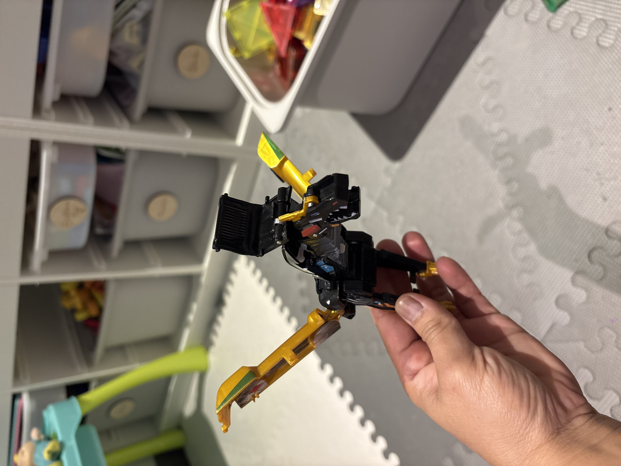 A person's hand is holding a black and yellow toy robot with wings. The toy is positioned in the foreground, with its wings extended. In the background, there is a white shelving unit filled with storage bins. Some bins contain colorful toys. The floor is covered with a light gray interlocking mat. The overall scene appears to be an indoor play area or toy storage space. The image does not contain any information that would indicate its location in Dubai, United Arab Emirates.